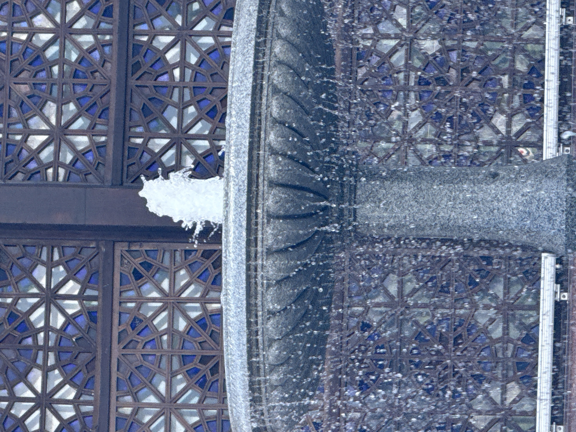 The image shows a close-up of a fountain with water cascading down a textured surface, likely stone or concrete. Behind the fountain, there are decorative screens or windows featuring a geometric pattern with dark brown framing and blue and white geometric shapes. These screens resemble those found in traditional architecture, possibly Islamic or Moorish in style. The water spray from the fountain creates a dynamic element, with numerous droplets visible against the darker background. The overall impression is one of a blend of natural elements (water) with intricate architectural design. The lighting suggests it could be daytime, but there are no clear shadows indicating a specific time. The presence of what appears to be ice or frost on the upper edge of the frame suggests cold weather conditions, consistent with a Moscow, Russia location. No people are visible in the image. The focus is on the texture of the fountain and the patterned screens, with the water creating a sense of movement and a somewhat abstract aesthetic.
