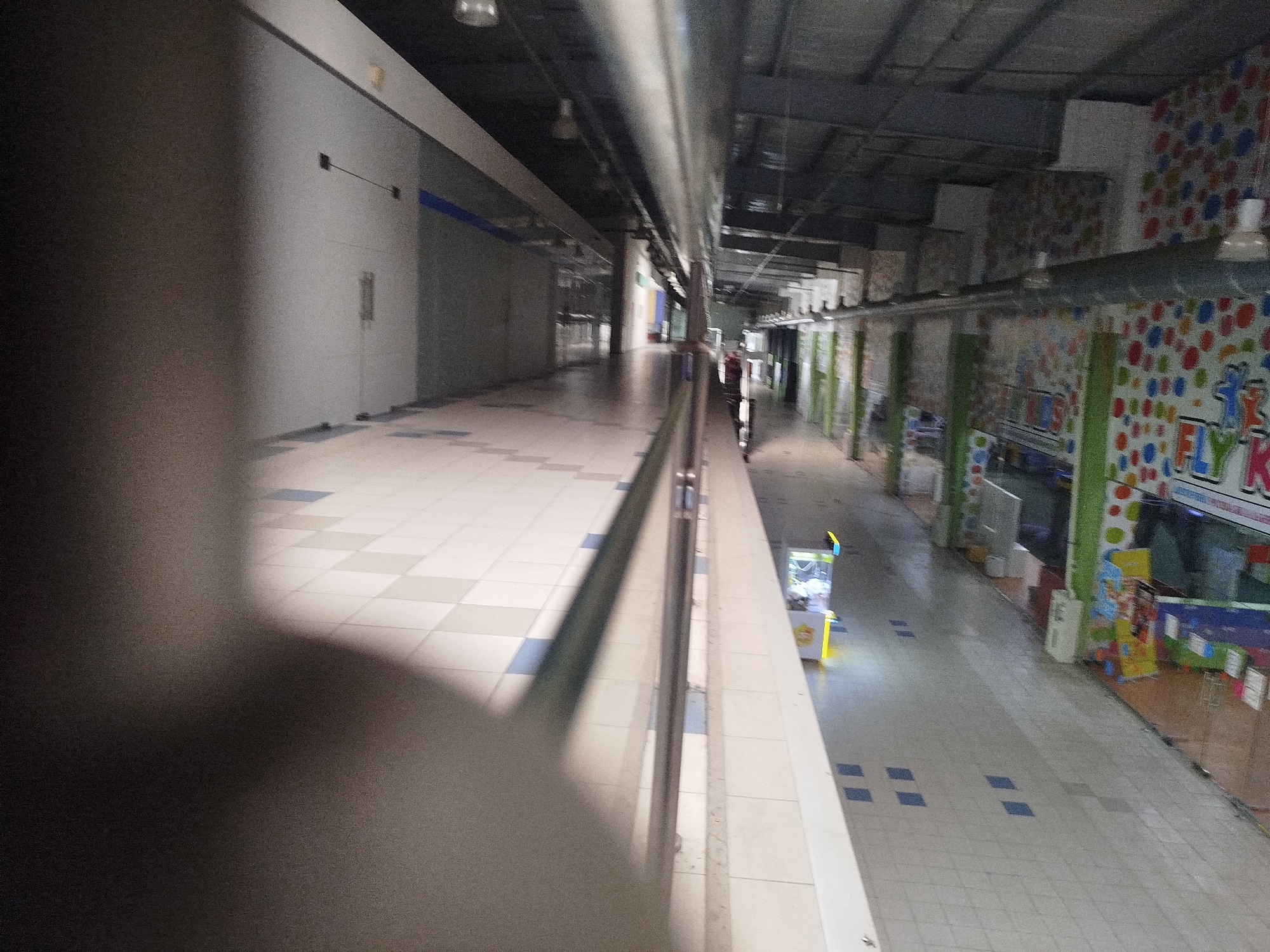 This image shows an interior view of a shopping mall in Kremenchuk, Ukraine. The perspective is from an elevated walkway or balcony, looking down a long, empty corridor. The floor is tiled with a pattern of light and dark squares, some of which are blue.
On the left side of the corridor, there are plain white walls, some with blue accents. Further down on the right, there are storefronts with brightly colored, polka-dot patterned walls, suggesting a children's play area or entertainment venue. Some signage with the letters "FLY K" is visible on these storefronts.
There are various objects visible along the corridor, including what appear to be trash receptacles, display stands, and possibly game machines. The lighting appears to be from overhead fixtures, and the overall atmosphere is dim and deserted. A single person, dressed in dark clothing, is visible walking down the corridor in the distance on the right side. The ceiling structure of the mall, with exposed beams and lighting fixtures, is visible in the upper portion of the image.