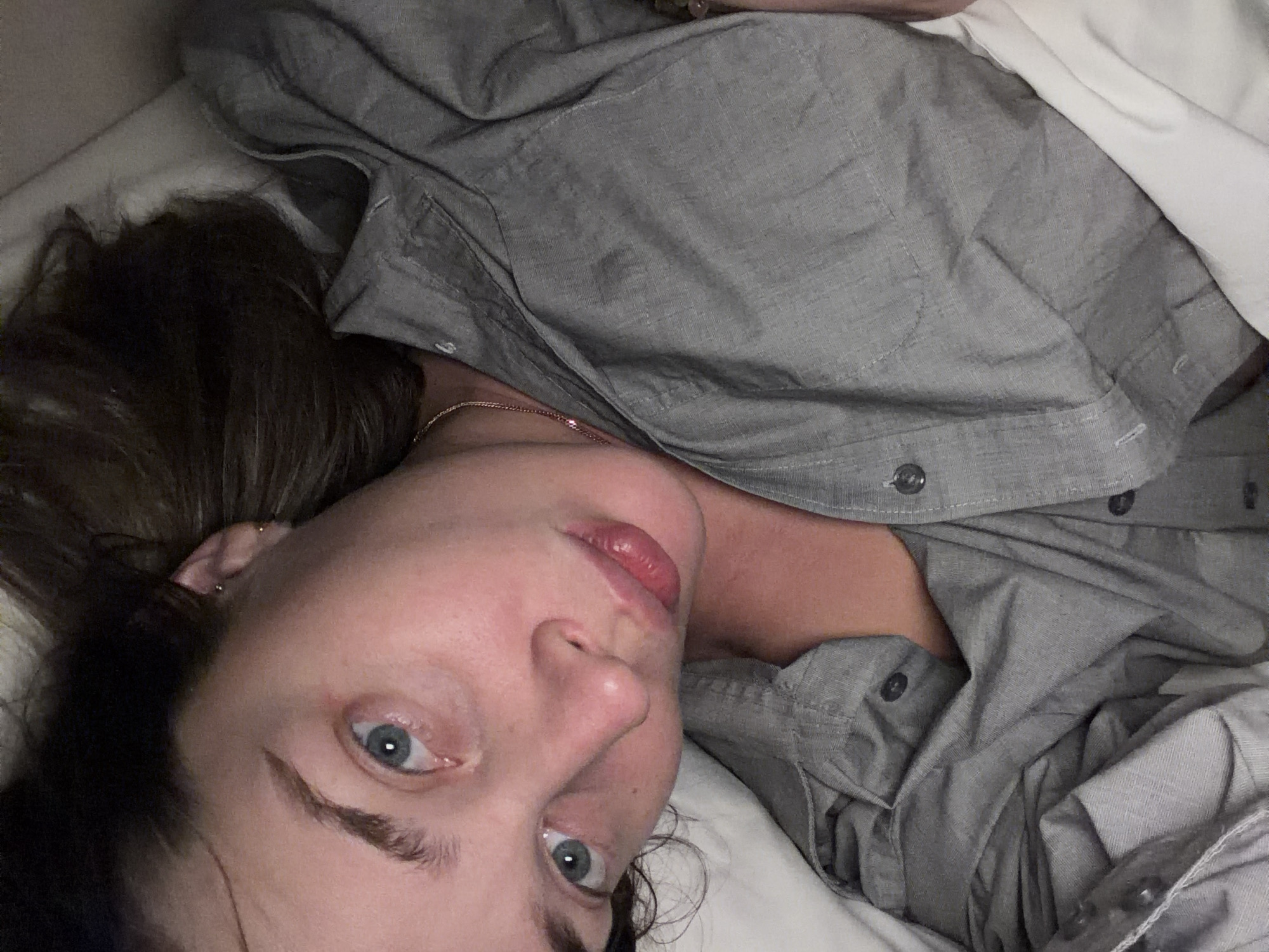 A woman is lying down, possibly in bed, wearing a gray button-down shirt. Her head is resting on a white pillow or sheet. She has blue eyes, light skin, and dark hair. She is looking directly at the camera. A gold chain is visible around her neck. The background is out of focus. There is no indication this photo was taken in Moscow, Russia.