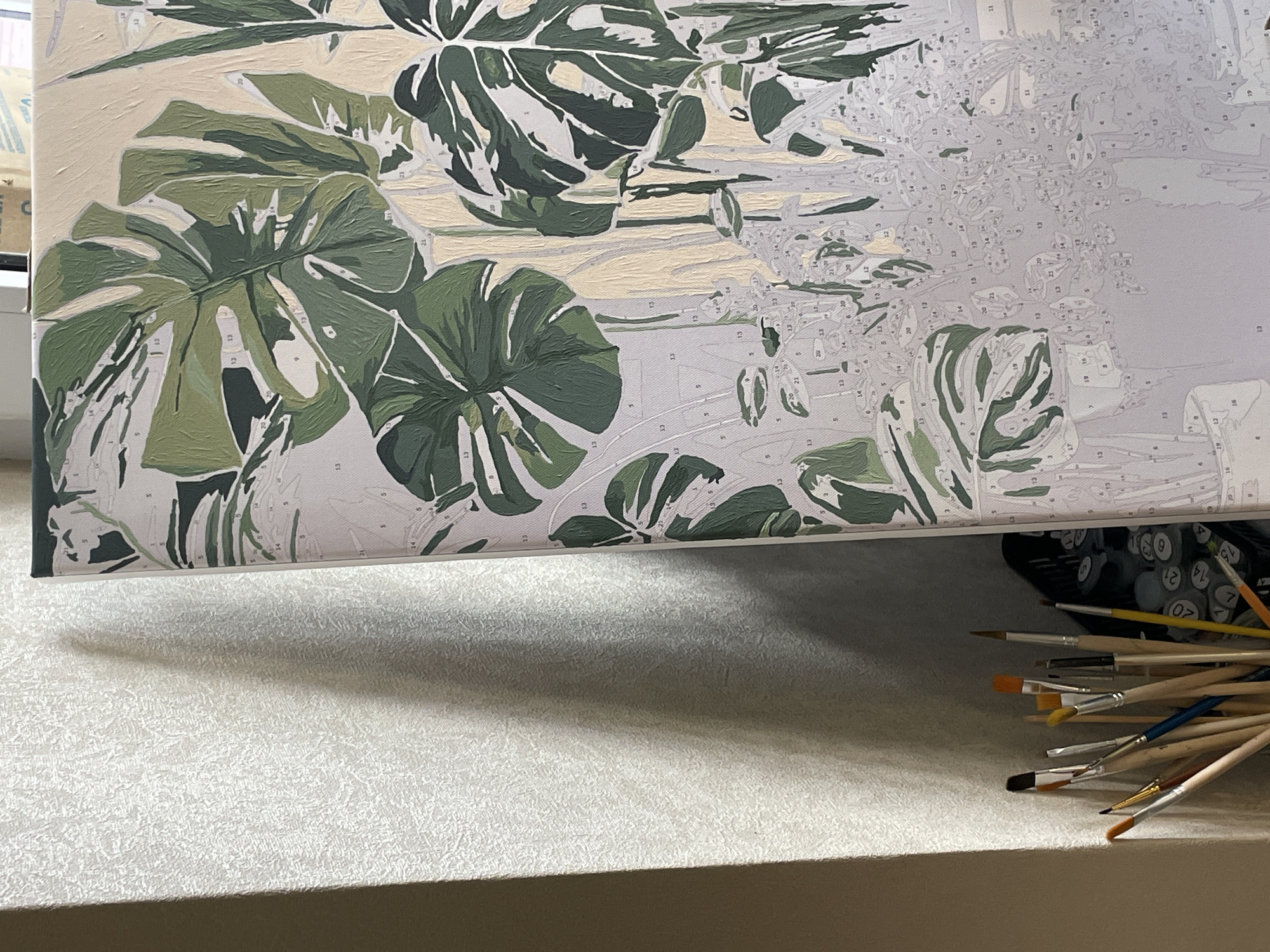 The image shows a close-up of a paint-by-numbers canvas depicting large green monstera leaves and other foliage. The canvas is partially completed, with many numbered sections filled in with various shades of green and beige. To the left of the canvas, a collection of paintbrushes in different sizes stands in a holder. The background to the left features a textured wall with dramatic shadows cast by light from an unseen source, suggesting interior lighting. The scene is likely set indoors, possibly in a home or studio. There is no indication of the city of Anapa, Russia, or any specific geographical location within the image itself.