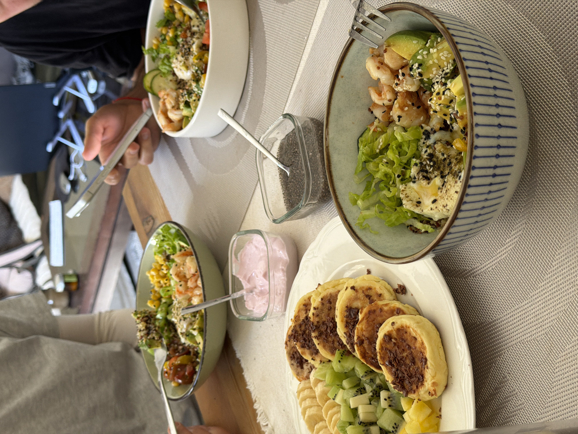 The image captures an indoor dining scene, likely in a residential setting, featuring a meal prepared for multiple individuals. A light-colored, patterned placemat covers a wooden table.
Three meal settings are visible. On the left, a person wearing a light grey t-shirt over a long-sleeved undershirt is shown holding a fork, poised above a light green bowl. This bowl contains a substantial salad, identifiable elements including shrimp, diced avocado, lettuce, corn kernels, quinoa, and chopped tomatoes.
In the foreground, a white plate holds several round, golden-brown pancakes or fritters, each topped with dark chocolate shavings. Arranged alongside the pancakes are slices of banana and diced kiwi fruit. Adjacent to this plate, two small, clear square glass containers are present: one holds pink yogurt with a small silver spoon, and the other contains black chia seeds, also with a small silver spoon.
In the foreground-right, a distinct patterned bowl (white with blue vertical stripes) contains another salad, featuring green lettuce, cooked shrimp, diced avocado, a white protein (possibly a poached egg), and corn, garnished with sesame seeds and dark seasonings. A fork rests inside this bowl.
On the right side of the frame, a person wearing a black t-shirt holds a silver smartphone vertically in their right hand, appearing to be actively using the device. A red bracelet is visible on this person's wrist.
In the blurred background, a white bowl containing similar salad components is visible. Further back, out-of-focus elements include a dark laptop resting on a stand and white charging cables on a darker surface, indicating a modern interior.
There are no discernible visual cues within the image that specifically identify the location as Tel-Aviv, Israel.