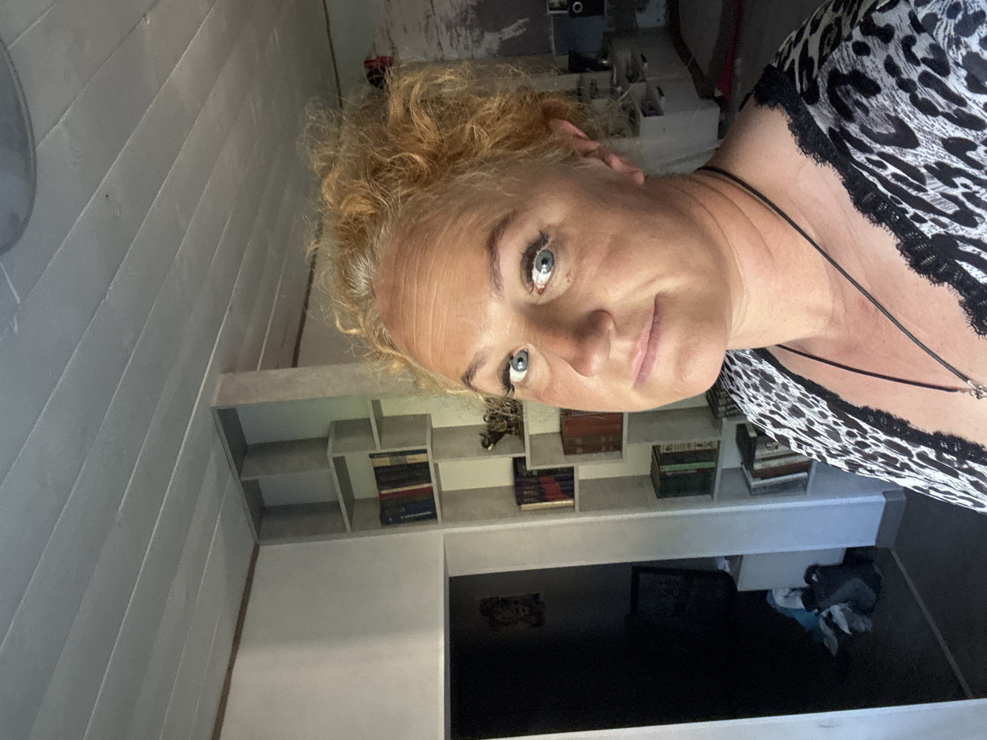 The image shows a woman with curly blonde hair and blue eyes taking a selfie in what appears to be a room. She is wearing a black and white animal print top with black lace trim. The background includes a white ceiling with horizontal planks, a white wall, and a tall, white shelving unit filled with books. To the left, there is a doorway leading to a dark room with some items on the floor. The overall impression is of an indoor, possibly domestic setting. The location in Chaikovsky, Russia, is not discernible from the visual information provided in the image.
