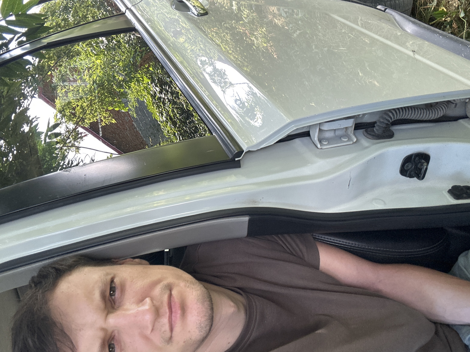 A man is sitting in the driver's seat of a white car, looking towards the camera. He has brown hair, a light complexion, and is wearing a brown t-shirt. The car door is open, revealing the interior and the exterior of the car. Outside the car, green trees and a brick building are visible. The photo appears to have been taken in Makiv, Ukraine.