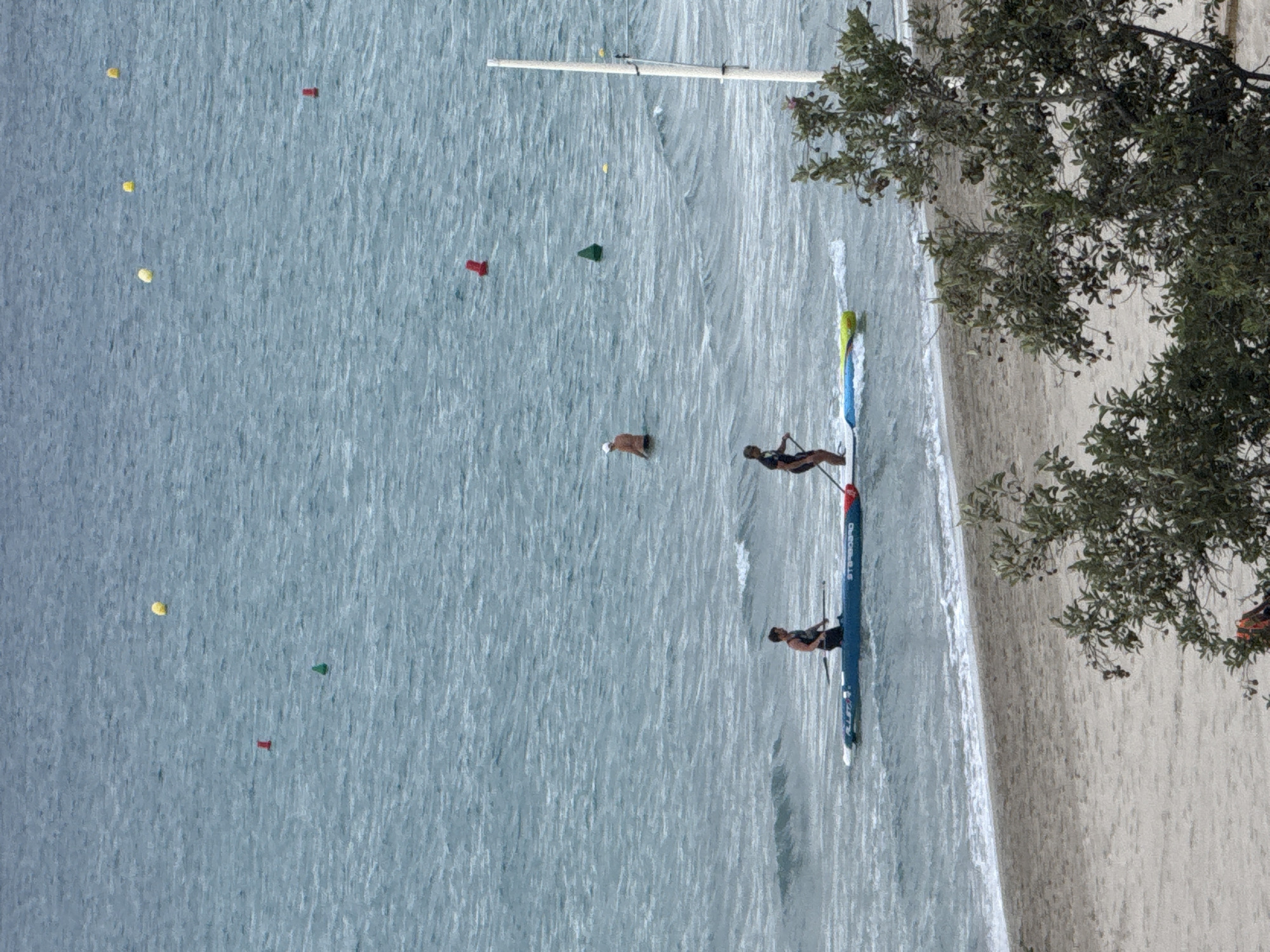 An aerial view captures a vibrant beach scene on a clear, sunny day, with the image rotated 90 degrees clockwise, making the water appear at the bottom and the beach and trees at the top right when viewed normally.
In the expanse of light blue-green water, which shows subtle ripples, three individuals are engaged in water activities. Two people are on paddleboards closer to the shore. One person, wearing dark swimwear, stands on a blue paddleboard with a yellow and blue tip, actively paddling. The text "STARBOARD" is visible on this board. Further to the left, another person in dark swimwear is positioned on a fully blue paddleboard, appearing to kneel or sit, with a paddle in hand. The words "STARBOARD PRO" and "ALLSTAR" are clearly discernible on this board. A third individual, wearing a light-colored cap or head covering, is floating or swimming further out in the water.
Scattered across the water are several colorful buoys: multiple yellow ones, red rectangular ones, and green triangular ones, suggesting a marked course or swimming area. A long, slender white pole or mast extends vertically from the water towards the top of the frame.
To the right, a sandy beach transitions into a lush green tree with dense foliage, providing shade. The overall atmosphere suggests a leisurely and active day at a coastal or lakeside location.
