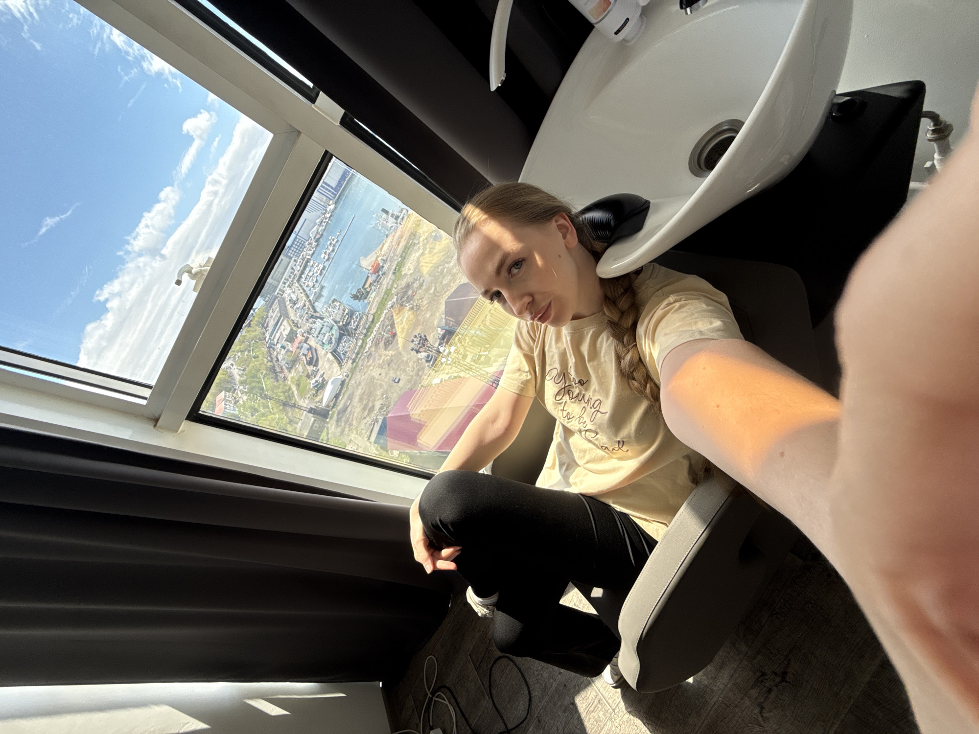 A young woman is captured in a selfie taken from a low angle, seated in what appears to be a hair salon, with a large window behind her providing a panoramic view. She has blonde hair tied in a braid and is wearing a light yellow t-shirt and black pants. Her head is positioned near a white, ceramic salon shampoo basin, which is integrated into a dark-colored chair unit. The woman looks directly at the camera with a neutral expression.
Bright sunlight streams through the window, illuminating the scene. Outside, a clear blue sky with scattered white clouds is visible. The window overlooks a vast landscape featuring a significant body of water, likely a bay or harbor, teeming with numerous boats, ships, and maritime infrastructure, consistent with the port city of Petropavlovsk-Kamchatsky, Russia. On land, there are buildings, industrial structures, and patches of green vegetation. Dark gray or black blinds are partially drawn along the bottom of the window frame.
On the woman's t-shirt, the partial text "Too Young to be Cool" is discernible. A power cord is visible on the light-colored wooden floor to her left. The overall impression is a casual moment in a salon setting with a striking urban and maritime backdrop.