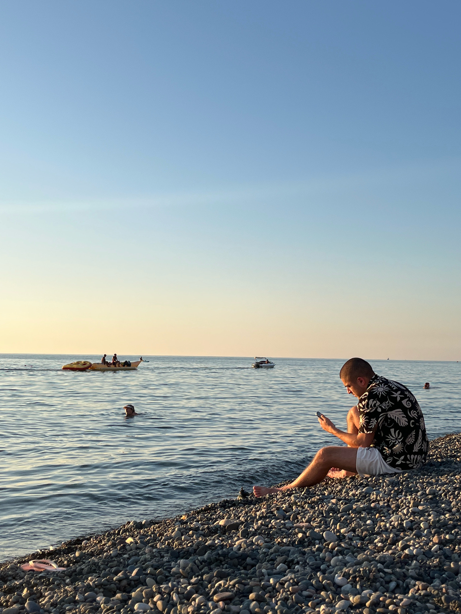 An individual wearing a black and white patterned short-sleeved shirt and white shorts is sitting on a pebble beach, looking down at their phone. Their legs are crossed, and their feet are bare. The sea stretches out behind them, with gentle waves lapping at the shore. In the distance, a banana boat with several people on it is being pulled by a boat, and another small motorboat is visible on the water. One person is swimming in the sea closer to the shore. The sky is a clear, pale blue, transitioning to a soft yellow hue near the horizon, suggesting either sunrise or sunset. A pink flip-flop is lying on the pebbles near the water's edge. The scene appears to be taking place in Kobuleti, Georgia.
