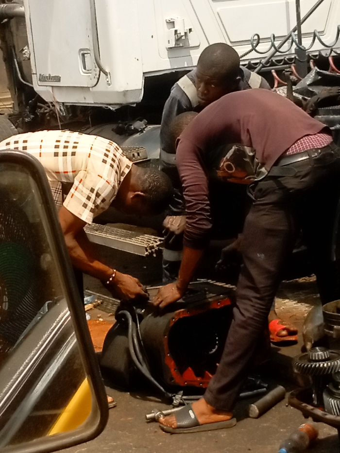The scene depicts three adult males engaged in vehicle repair work outdoors in Ogidi, Nigeria. A large, dark-colored mechanical component, likely a transmission or differential housing with a red-painted interior rim, lies on a stained ground surface. Scattered around it are various tools and disassembled metallic gear components. In the background, part of a white truck cabin, bearing a partially visible "MACK" emblem, is present.
To the left, one man, wearing a beige and brown checkered short-sleeved shirt and a beaded bracelet on his right wrist, is bent forward, actively manipulating the mechanical component with both hands. To the right, another man, dressed in a long-sleeved maroon shirt, dark trousers, and grey slide sandals, is similarly bent over and focused on the component. Between these two, slightly behind and observing, stands a third man in a dark blue work uniform. A black side-view mirror frame partially obstructs the immediate left foreground, reflecting a dark, cylindrical object. A red slipper and a small plastic bottle are visible on the ground.