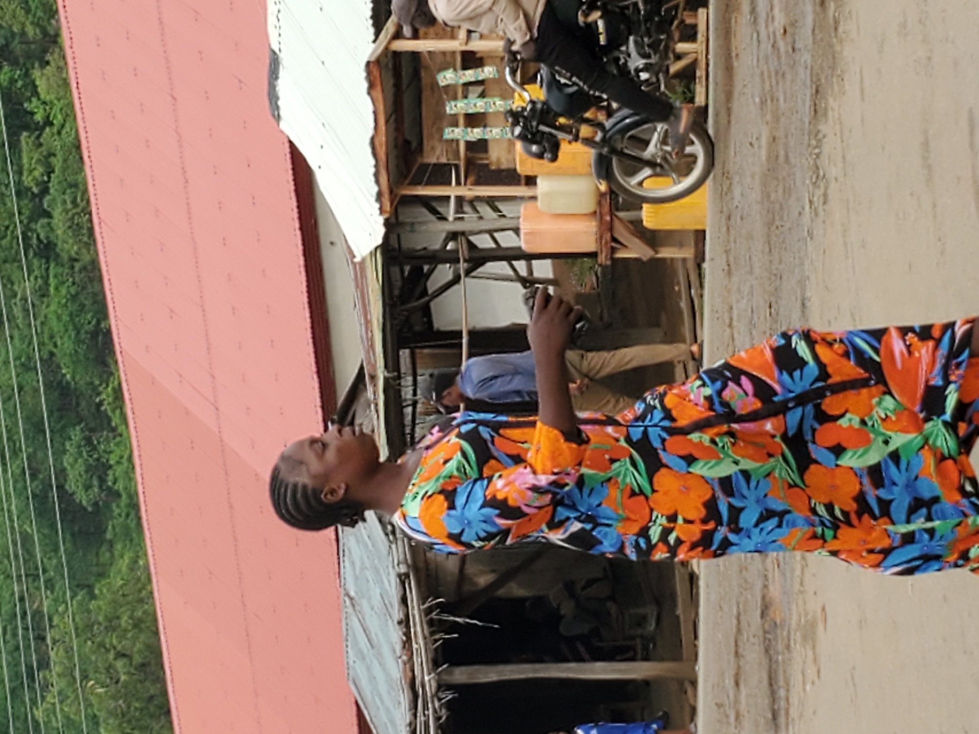 The image captures a street scene in what appears to be Mararaba, Nigeria. A woman wearing a brightly colored floral dress with blue, orange, and black patterns is the focal point. She is facing left with her head tilted back, seemingly looking at something above. Her hair is styled in cornrows.
Behind her are partially visible structures: a building with a red roof to the left and a makeshift shop with a white roof in the background. The shop seems to sell goods, with price tags hanging. A man is sitting on a motorcycle in front of the shop. Another man is standing nearby. Several plastic containers of varying colors are visible in the shop's framework. The street surface is a light, rough texture. Lush green foliage is visible in the background.