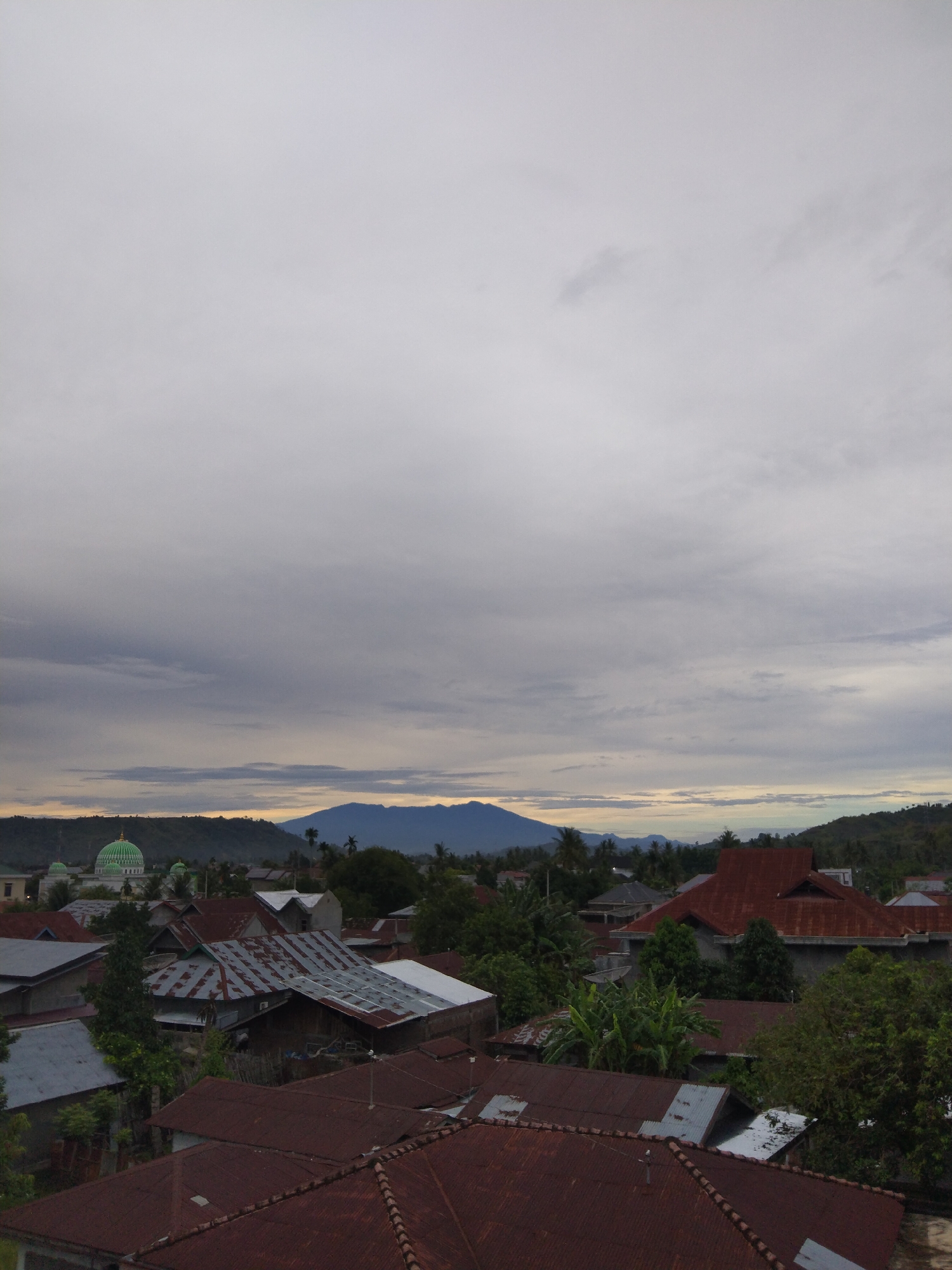 The image shows an aerial view of Bireuenmeunasahdayah, Indonesia, under an overcast sky. The foreground is dominated by a dense collection of rooftops, predominantly terracotta and rusted corrugated iron. Scattered trees are interspersed among the buildings. In the midground, a mosque with a green dome stands out against a backdrop of low hills. Further in the distance, a mountain range is visible on the horizon.