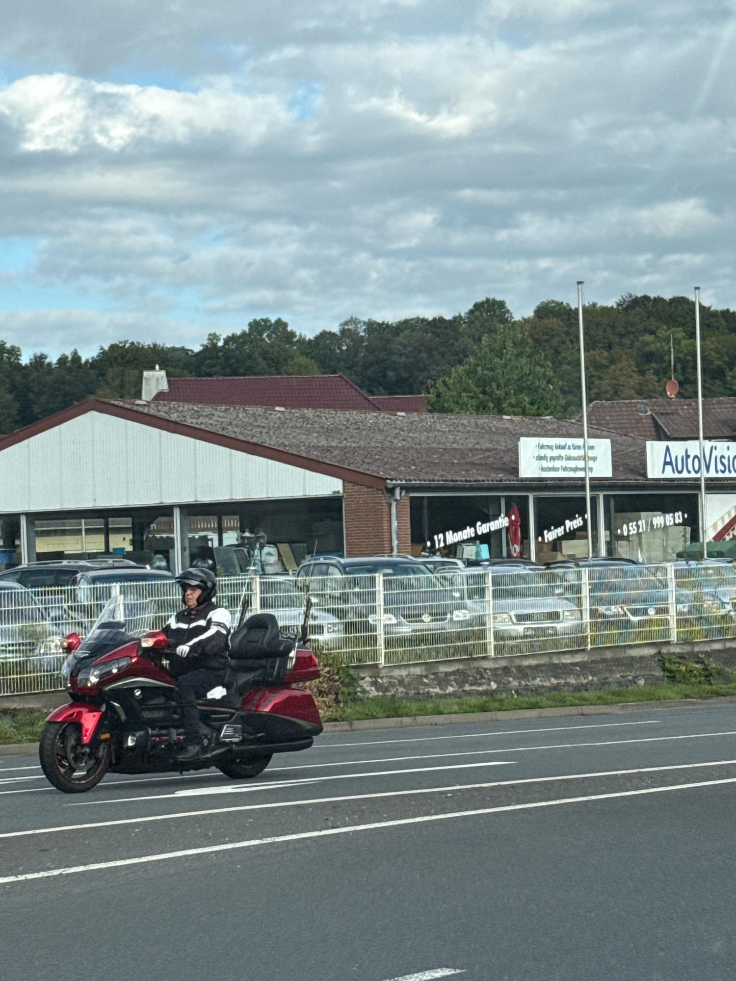 A motorcyclist wearing a black and white jacket and a helmet rides a red motorcycle on a road in Herzberg, Germany. The motorcycle has a large windshield and a passenger seat with a backrest. Behind the motorcycle is a car dealership with a building featuring large windows. Several cars are parked behind a fence in front of the dealership. The sky is cloudy.