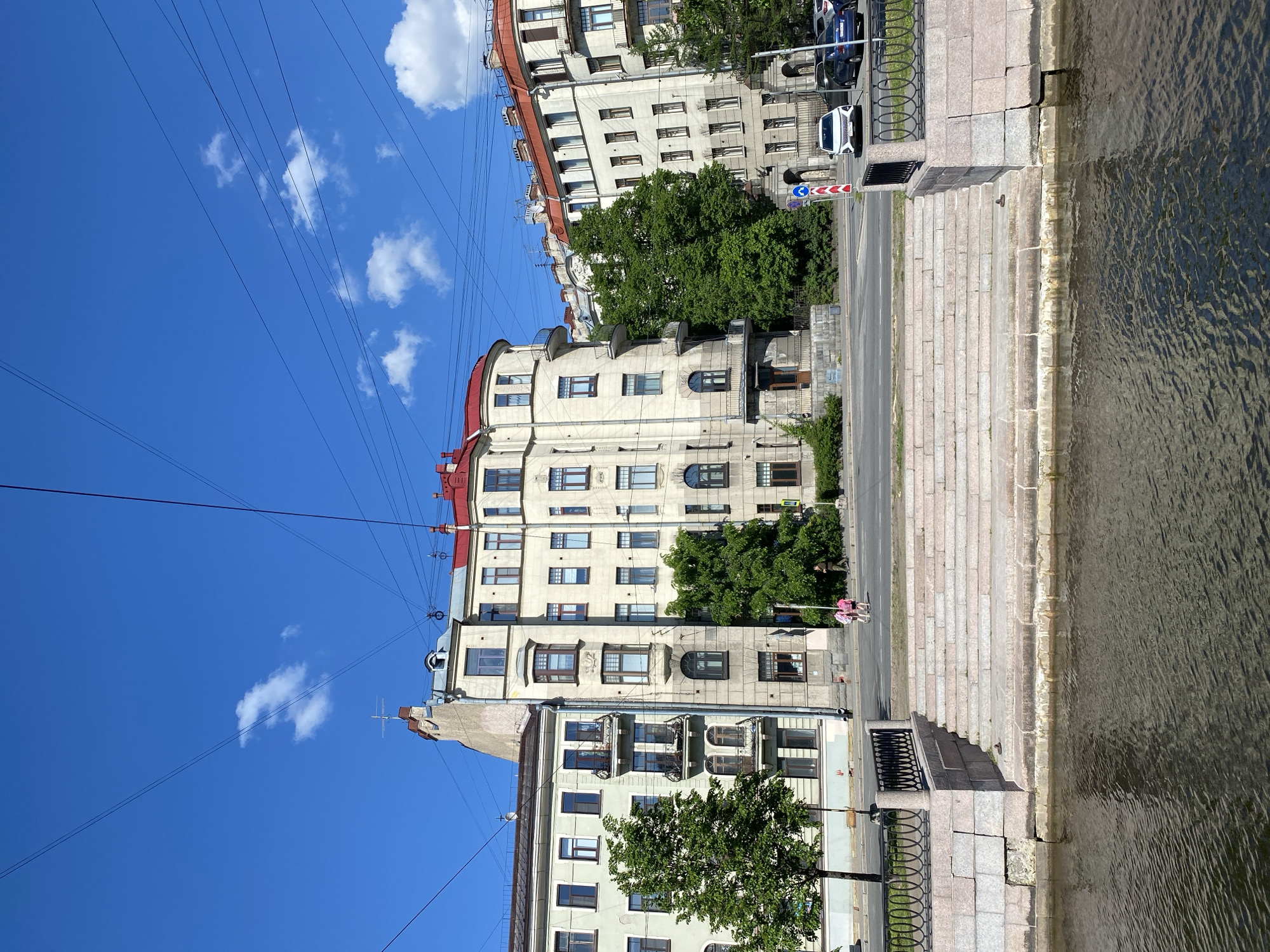 The image captures a sunlit urban waterside scene in Saint Petersburg, Russia, showcasing a canal or river on the right, bordered by a street and historic buildings on the left. The sky is a vibrant blue, adorned with scattered white cumulus clouds and crisscrossed by numerous black overhead power lines.
On the left, several multi-story buildings with light-colored facades, varied window shapes (including arched ones), and red or brown sloped roofs line the street. Some feature small balconies, a few of which are enclosed, and subtle architectural details. Green trees are interspersed along the street, casting shadows.
To the right, a wide granite embankment with steps descends to the calm, rippling water of the canal. A black, ornate metal railing runs along a section of the embankment. Adjacent to the embankment is a paved street with parked and moving vehicles, including a white car and a dark blue car. A blue, round traffic sign indicating a sharp right turn, along with a red-and-white chevron sign, is visible beside the road. A person wearing a pink top is seen riding a small bicycle on the street or sidewalk.
The scene depicts a clear, sunny day, likely morning or early afternoon, with strong natural light. The overall atmosphere is tranquil, portraying typical urban activity. The distinctive architecture, canals, and granite embankments are strong visual cues identifying the location as Saint Petersburg. No explicit text is readable on the buildings or vehicles, only the symbols on the traffic signs.
