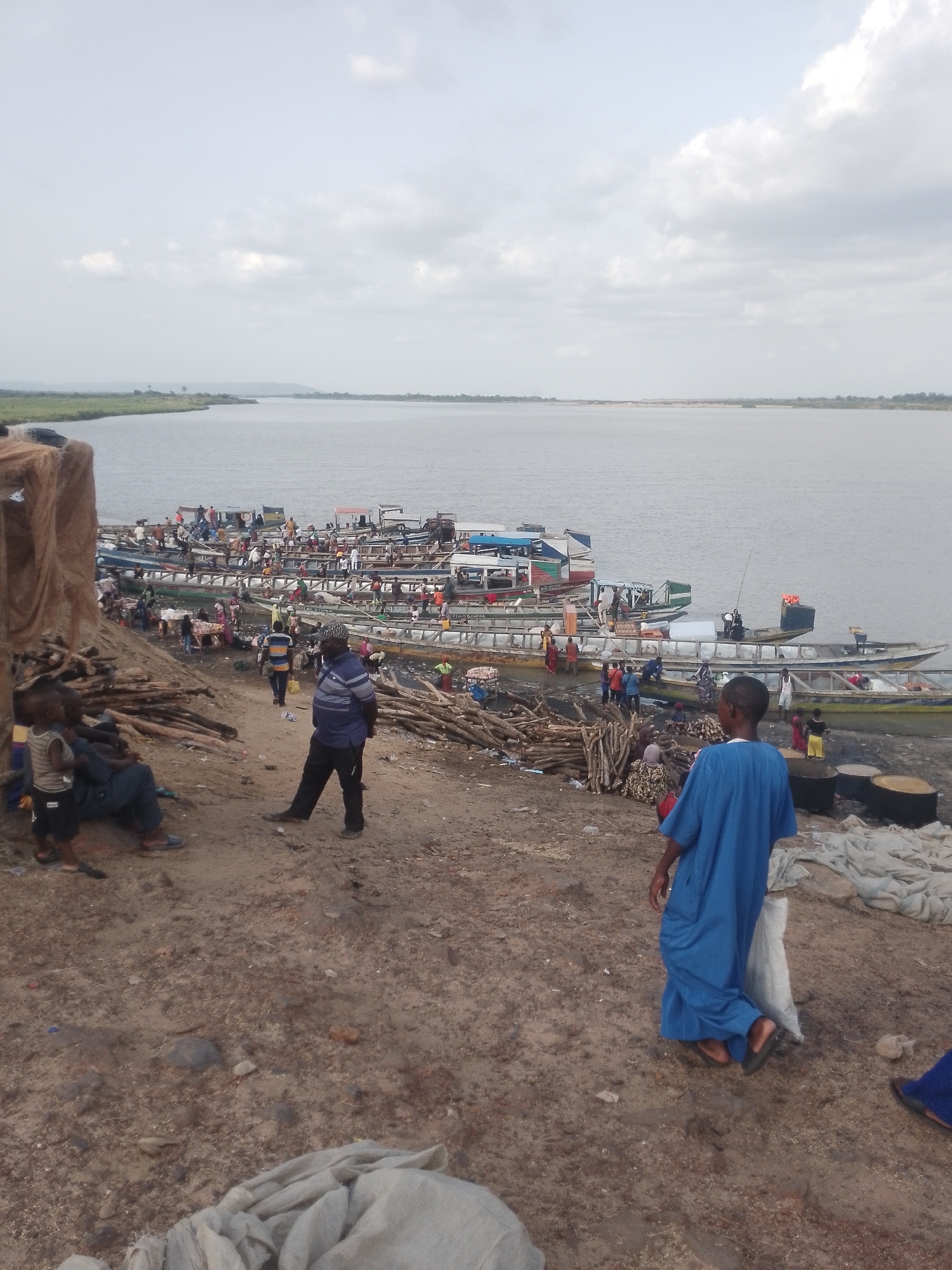 The image depicts a crowded riverfront scene, likely a port or harbor, in an unidentified city in an unknown region. Numerous boats are moored along the riverbank. People are milling around on the shore. One person is wearing a blue robe, a few others can be seen in casual attire, and logs are piled on the riverbank. The water is calm, and the sky is overcast.