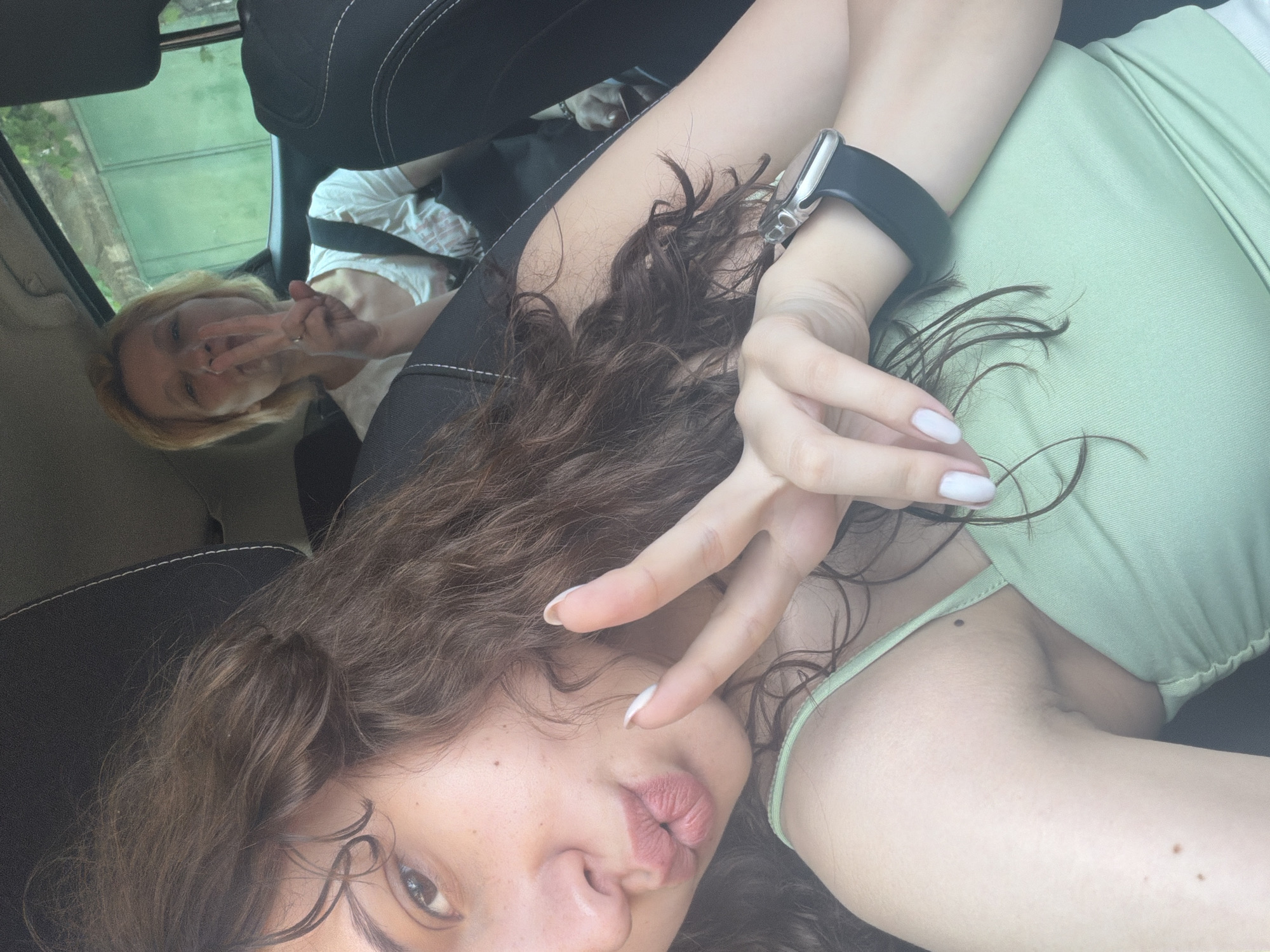 A playful selfie captures two women inside what appears to be a car. The image is taken from a slightly high angle, looking down towards the subjects.
In the foreground, a woman with dark, curly hair is prominent. Her head is tilted, showing one eye, her nose, and her lips pursed in a pouting or kissing expression. She is making a peace sign with her left hand, which is held close to her face. She wears a light green top, and a smartwatch with a black strap is visible on her left wrist. Her fingernails are painted white.
Behind and above her, another woman with blonde hair is visible, looking towards the camera. She is also making a peace sign with her hand and is wearing a white top. Both women appear relaxed and in good spirits, suggesting a friendly interaction.
The setting is an automobile interior, indicated by the dark car seats and headrests that frame the subjects. Through the car window in the upper left, green foliage and possibly some outdoor structures can be vaguely seen, suggesting it is daytime and they are either driving or parked. There is no visible text in the image.
