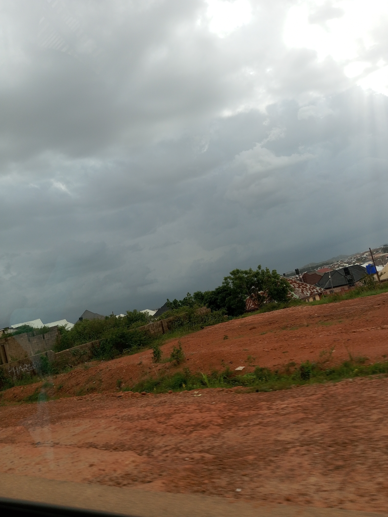 The image depicts a landscape with a cloudy sky and a view of a settlement. In the foreground, there is a reddish-brown dirt road or slope with some sparse green vegetation. A concrete wall with some graffiti is visible on the left side of the foreground slope. Beyond the slope, there are several buildings with various roof types, including corrugated metal and what appear to be tiled roofs. Dense green trees and bushes are interspersed among the buildings. In the background, a larger settlement or city is visible with more buildings extending into the distance, partially obscured by the overcast sky. The sky itself is filled with dark, heavy clouds, with shafts of light breaking through in the upper right portion of the image, suggesting either sunrise or sunset, or a break in the clouds. The overall impression is one of an urban or semi-urban area under a potentially stormy or overcast sky.
