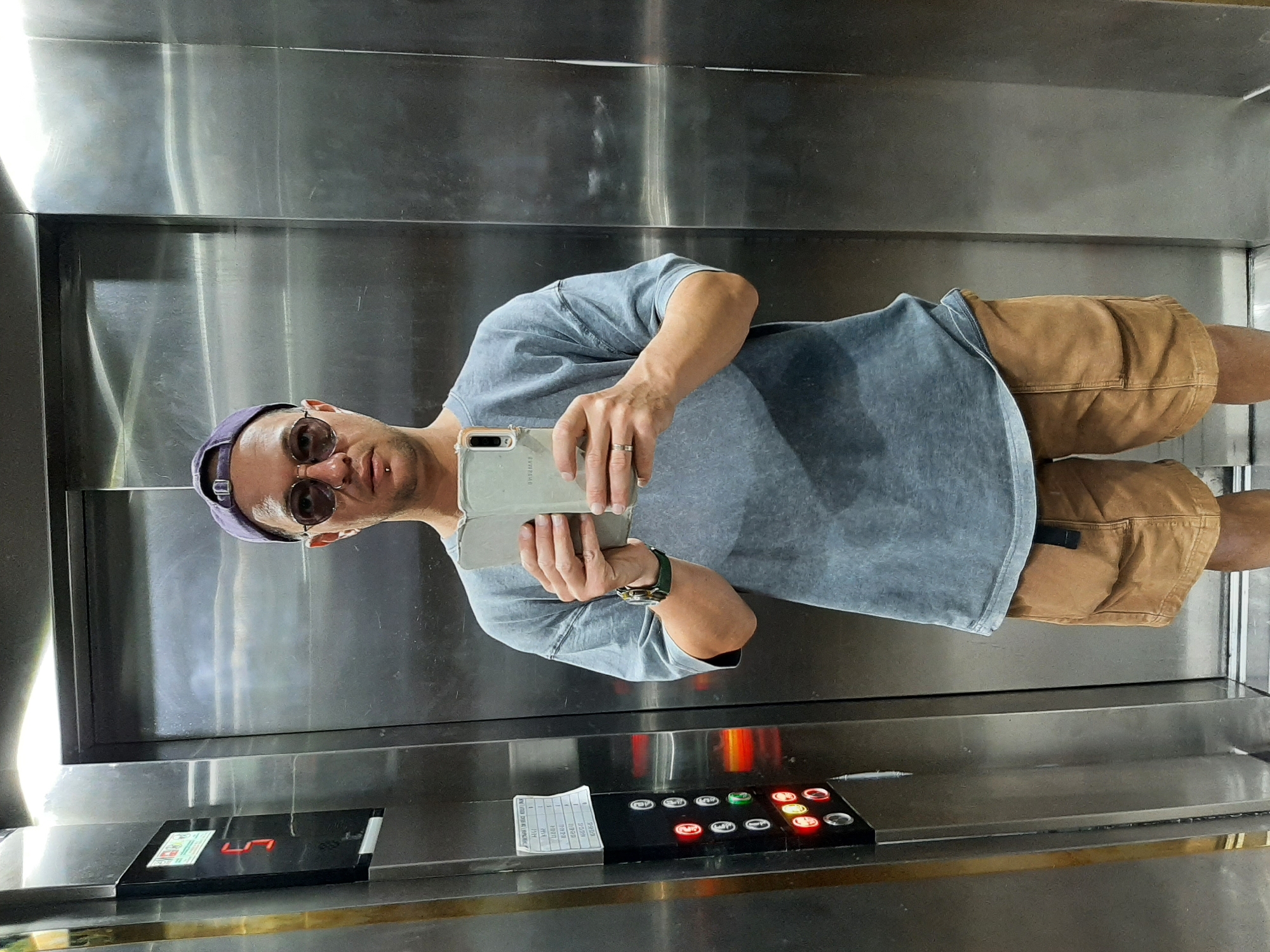 A man is taking a selfie in an elevator with stainless steel walls. He is wearing a grey t-shirt, tan shorts, sunglasses, and a purple baseball cap. He is holding a smartphone in his right hand. The elevator's control panel is visible and shows that the elevator is on floor "UT". A handwritten note is attached next to the buttons. Based on visual clues, it is possible that the elevator is located in a building in the city of Alanya, Turkey.