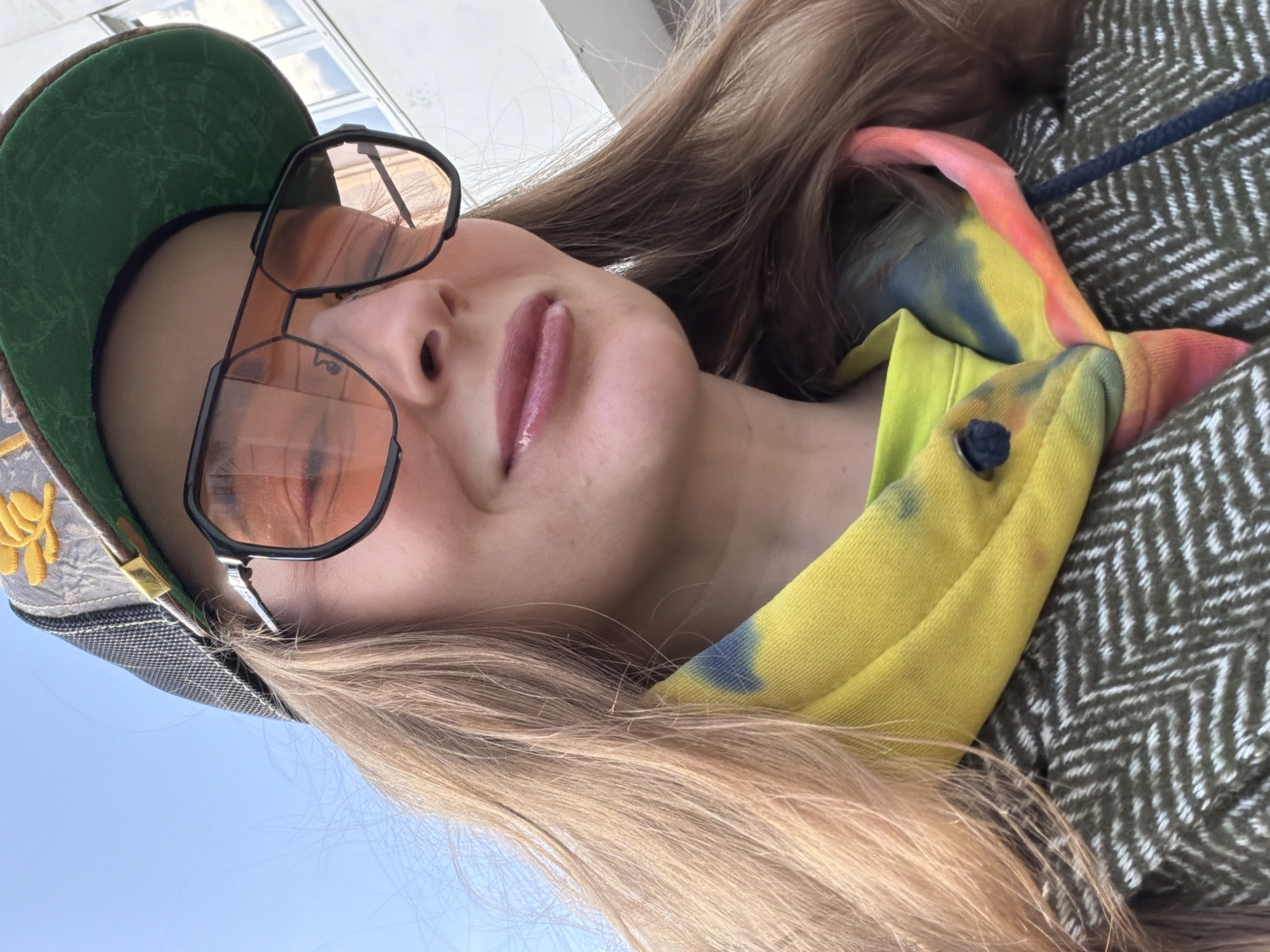 A close-up, low-angle self-portrait features a person with long, light brown hair, likely taken outdoors in Moscow, Russia during bright daylight. The subject is looking upwards at the camera with a neutral to slightly smiling expression. They wear large black-framed, aviator-style sunglasses with orange-tinted lenses, which reflect the clear blue sky and some architectural elements.
The person is wearing a green baseball cap with a black mesh brim. The cap features yellow embroidery on its front, which appears to be a stylized logo or letters, though not clearly discernible. Their lips are slightly parted and appear to have a light pink or nude gloss.
They are dressed in a bright yellow (possibly lime green) hoodie or sweatshirt that is vibrant with tie-dye patterns in blue, pink, and orange. A dark button or toggle is visible on the hoodie. Over this, they wear a thick outer jacket with a distinct green and white herringbone pattern. A dark blue cord or drawstring is visible near the jacket's collar.
The background primarily consists of a clear blue sky on the left side, indicating fair weather. On the upper right, reflections of windows or a building are visible, suggesting an urban or semi-urban setting. The bright lighting confirms it is daytime. No clearly legible text is visible other than the indistinct embroidery on the cap.
