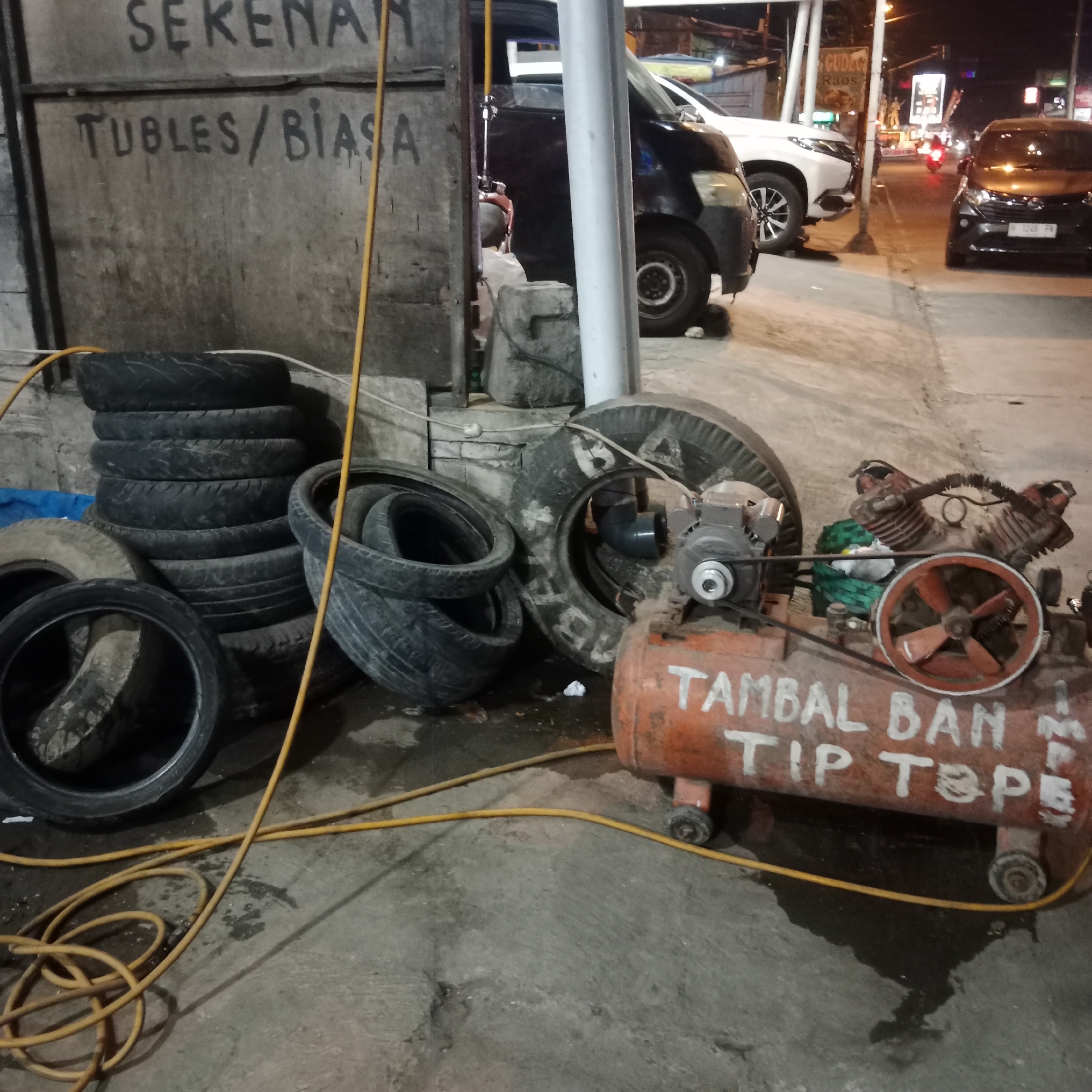 The image depicts a tire repair shop at night in Gubug, Indonesia. The shop appears to be located outdoors, possibly on a street or sidewalk, with vehicles visible in the background. The foreground is dominated by a collection of used tires stacked haphazardly, a yellow air hose coiled on the ground, and an orange air compressor. The air compressor has white lettering that reads "TAMBAL BAN TIP TEPE," which translates to "Tire Patch Tip Top." The wall behind the tires has handwritten text in Indonesian, including "SEKENAN" and "Tubles/Biasa," suggesting services for tubeless and regular tires. The lighting is dim, typical of nighttime, with artificial lights illuminating the scene. The ground is concrete, with some dampness and debris present. In the background, several cars are parked, and streetlights and building lights suggest a populated area. No people are clearly visible in the image.