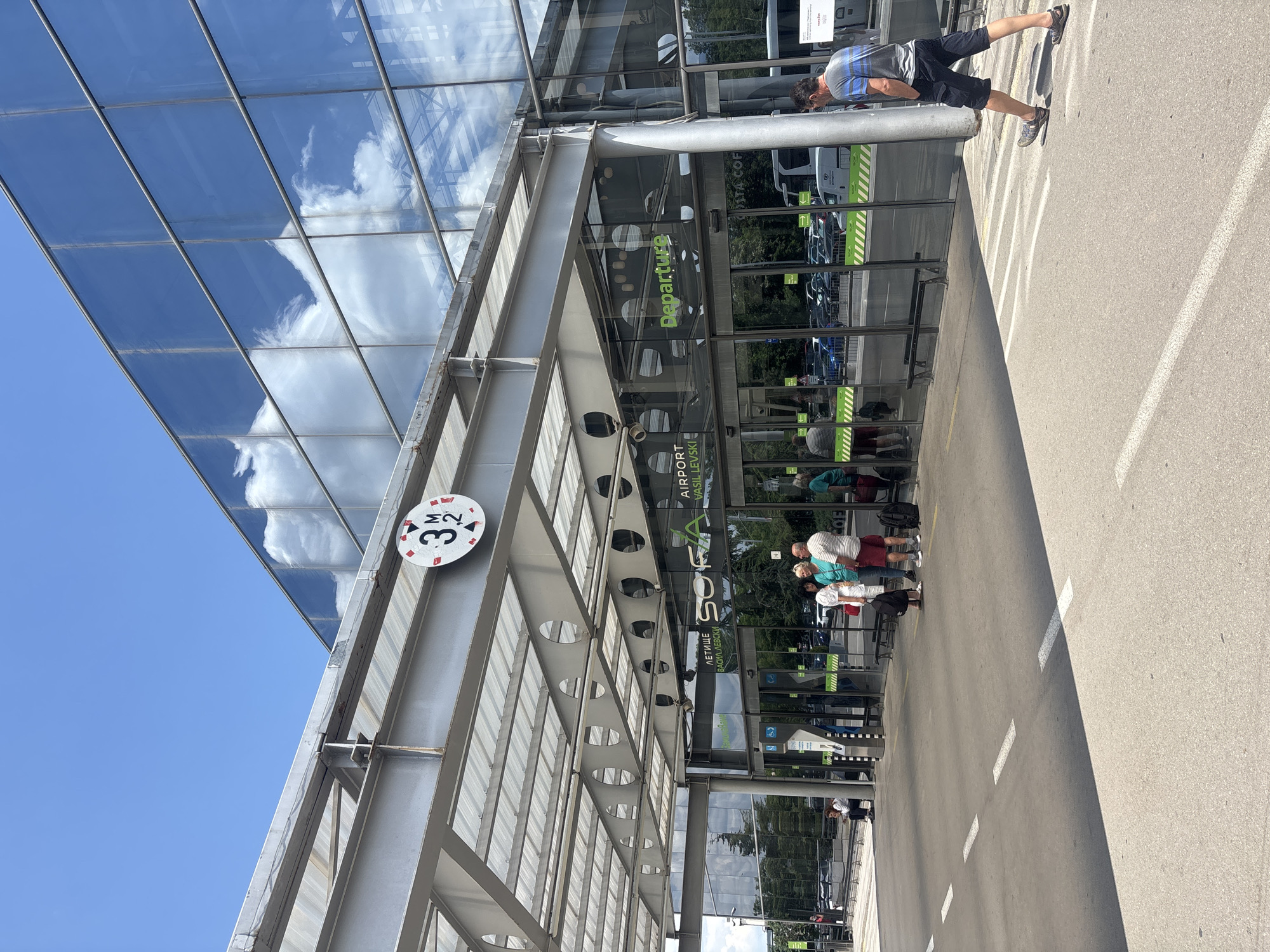 The image shows the exterior of Sofia Airport in Sofia, Bulgaria, on a clear, sunny day. The airport terminal features a modern glass facade reflecting the blue sky and clouds. Above the entrance, a circular sign indicates a height restriction of 3.2 meters. Several people are visible, some waiting outside the entrance, others walking away from the building. A man in a striped shirt and shorts walks towards the right side of the frame. Cars and a bus are parked in the background, suggesting a functional airport environment. The scene is well-lit and captures the daily activity at the airport.