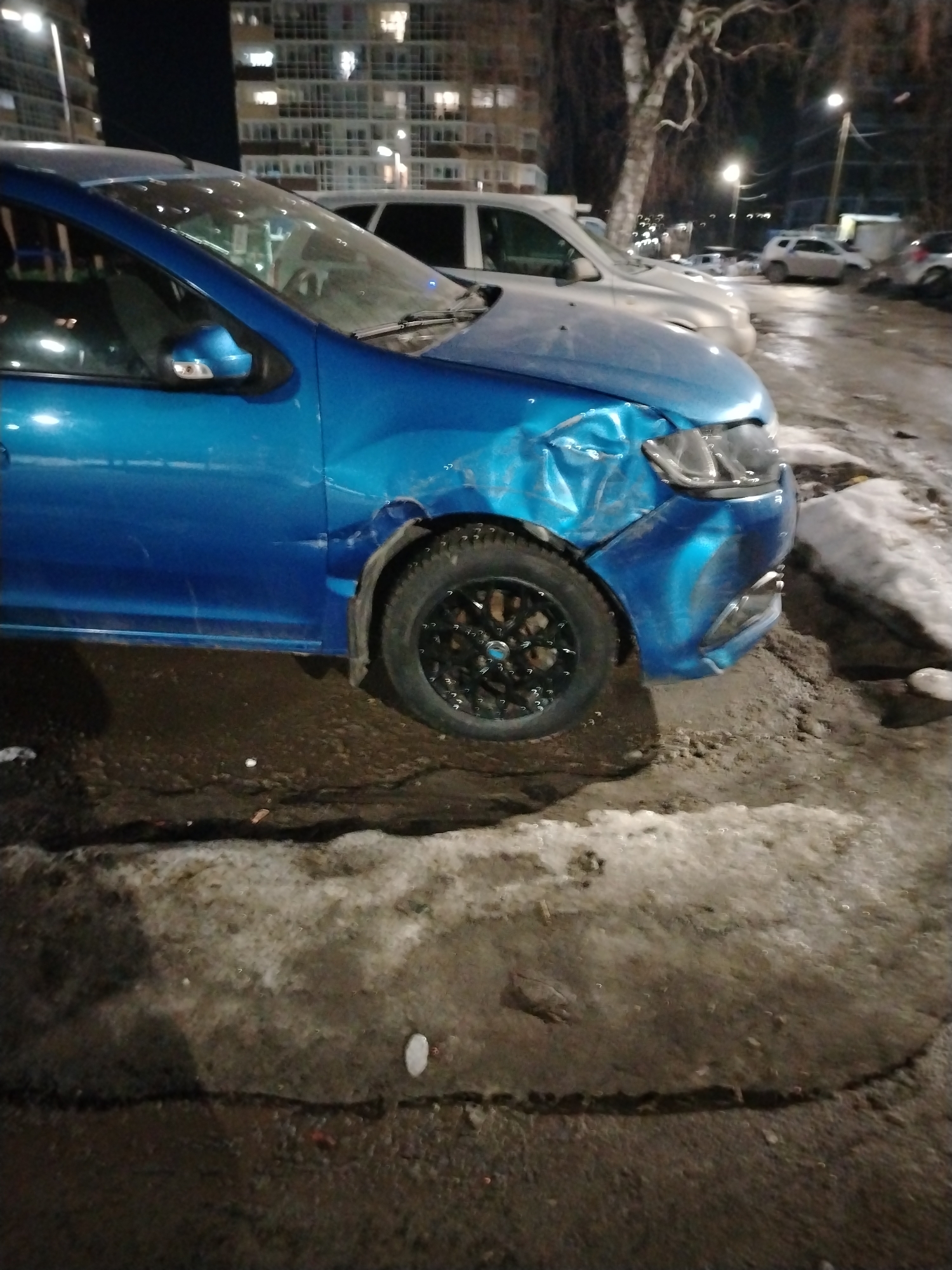 The image depicts a blue car with significant damage to its front passenger side. The fender is crumpled, and the headlight is damaged. The car's tire is black with a multi-spoke design. The car is parked on a road surface that appears to be wet and muddy, with patches of snow visible. In the background, there are several other cars, including a white SUV, parked along the street. Buildings with illuminated windows suggest an urban environment, possibly an apartment complex. Streetlights are also visible. Based on the location information, this scene is in the city of Izhevsk, Russia. There are no people visible in the image.