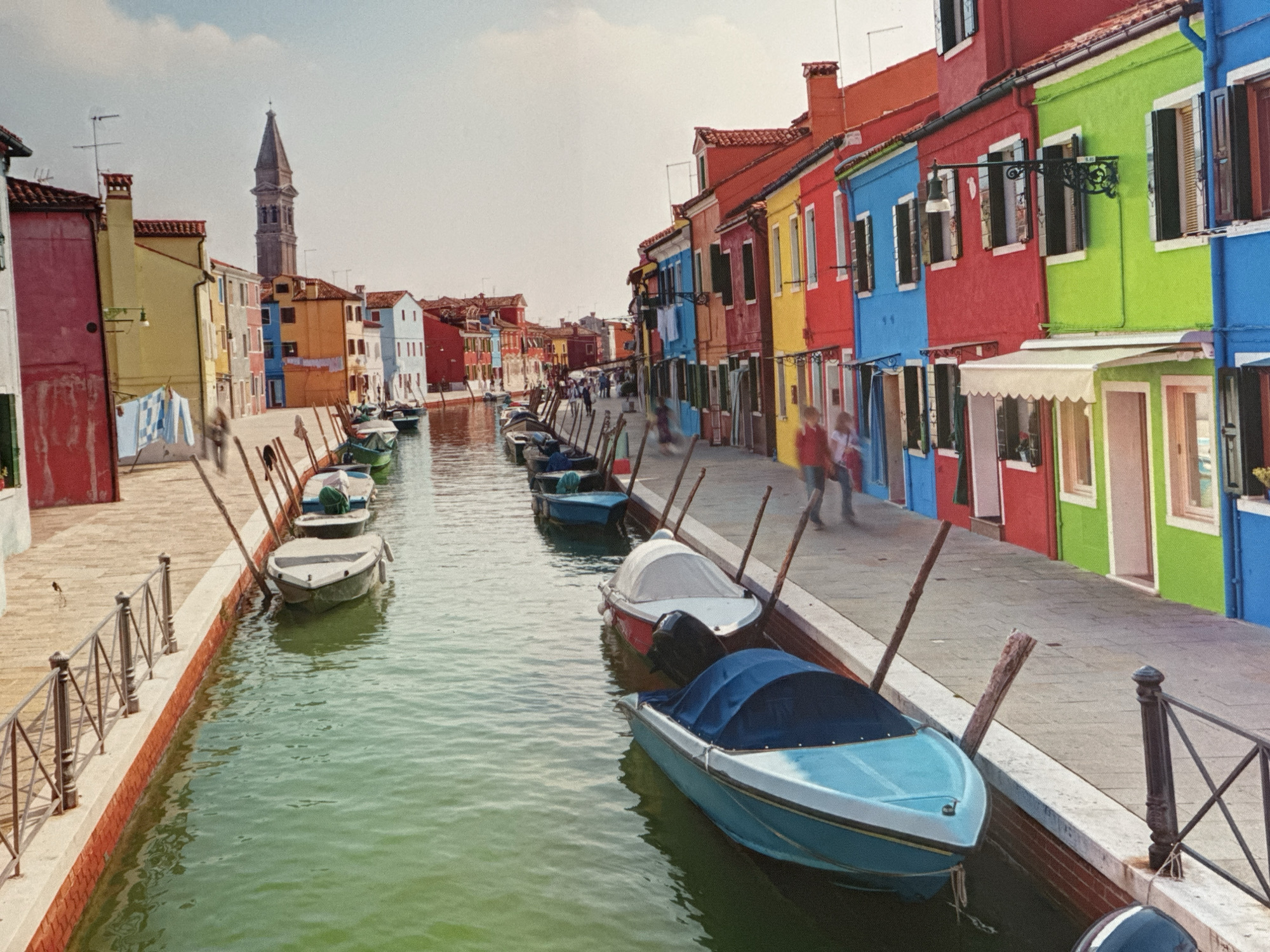 The image depicts a picturesque canal scene lined with brightly colored buildings and a row of parked boats. The architecture features houses painted in vibrant hues of pink, yellow, blue, red, and green, with traditional shutters and visible chimneys. A tall, slender bell tower with a pointed spire is visible in the distance, rising above the colorful rooftops.
Along the canal, several small boats are moored, with some covered and others open. The water of the canal is a greenish hue, reflecting the colorful surroundings. A paved walkway runs alongside the canal, with a decorative metal railing. A few indistinct figures of people can be seen walking along the walkway. The sky above is bright and mostly clear, with a few wispy clouds. The overall atmosphere is one of charming, lively urban scenery.
However, the initial premise of the location being Ossenzijl, Netherlands is incorrect. This scene is characteristic of Burano, an island in the Venetian Lagoon, Italy, known for its brightly colored houses and canals.