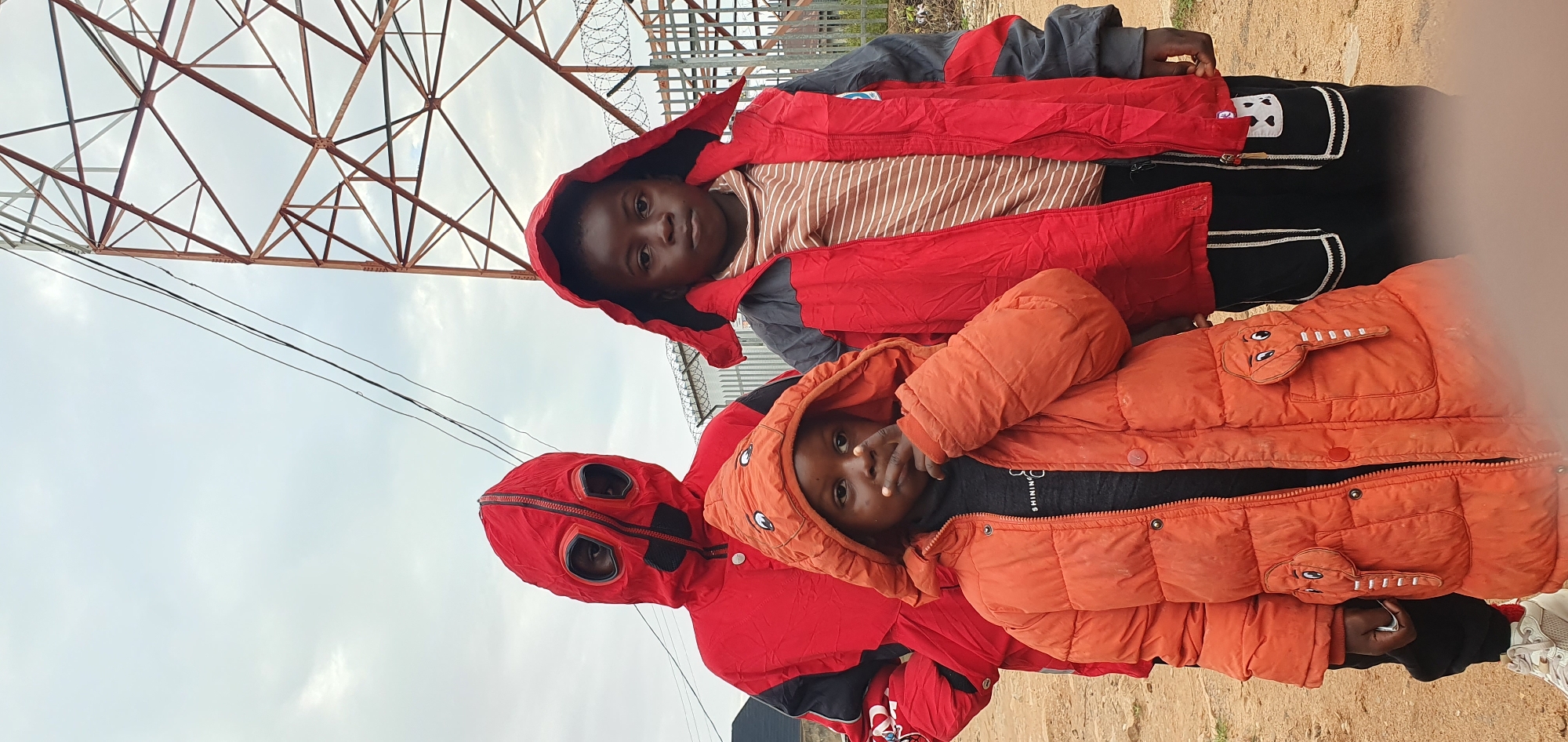 Three young individuals are standing outdoors in front of a metal tower structure. The person on the far left is wearing a striking red hooded jacket with black accents and a zippered front that resembles a mask with large circular eye openings. The middle person, a younger child, is in the foreground, wearing a bright orange puffer jacket with elephant-shaped pockets and a hood. This child is touching their mouth with their right hand. To the right, another individual wears a red and gray hooded jacket over a striped shirt. The ground appears to be dirt or gravel. In the background, a tall, lattice-like metal tower dominates the upper right portion of the frame, with power lines running across the sky. A metal fence with barbed wire is visible behind the individuals, suggesting a secured or industrial area. The sky is overcast and a muted gray. This scene is located in the city of Bukuru, Nigeria.