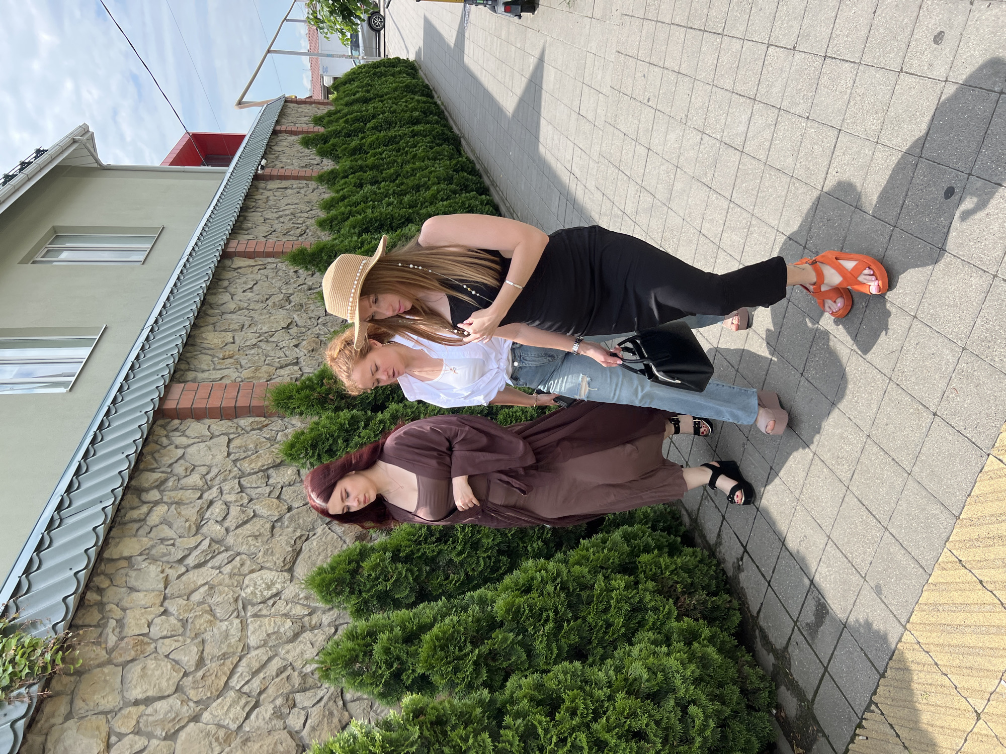 Three young women are walking on a paved sidewalk next to a stone wall and a line of evergreen trees. The woman on the left has long, dark red hair and wears a long, loose brown dress and black platform sandals. The woman in the middle has blonde hair and wears a white shirt, ripped blue jeans, and light pink platform sandals. The woman on the right wears a straw hat with pearl embellishments, a black sleeveless dress, and bright orange platform sandals. She is carrying a black handbag. The scene appears to be in an outdoor, urban setting, possibly a residential area or a commercial district, with a building visible in the background. The photo was taken in Krasnodar, Russia.
