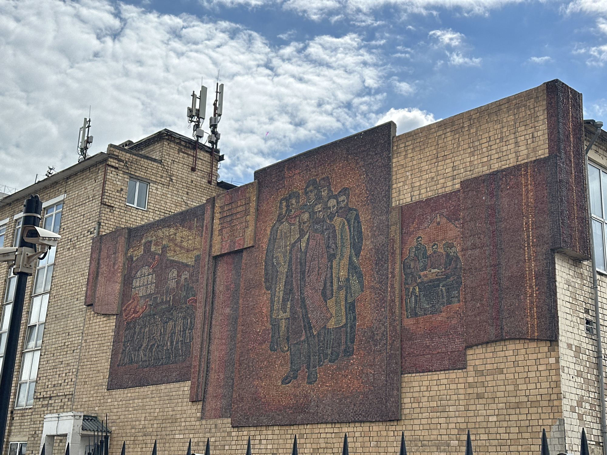 A low-angle, eye-level shot depicts a brick building under a partly cloudy sky. The building is adorned with three mosaic panels. From left to right, the panels show: a procession with a large building in the background, a group of figures in long coats and hats, and a group seated around a table.
On the roof of the building are a series of communication antennas. A security camera is mounted on a pole to the left of the image, and a wrought iron fence runs along the bottom of the frame. The building's construction, featuring yellow-toned bricks, is visible, with sections of the facade extending outward.
Based on the presence of Soviet-era imagery and other visual cues, it is located in Krasnoyarsk, Russia.