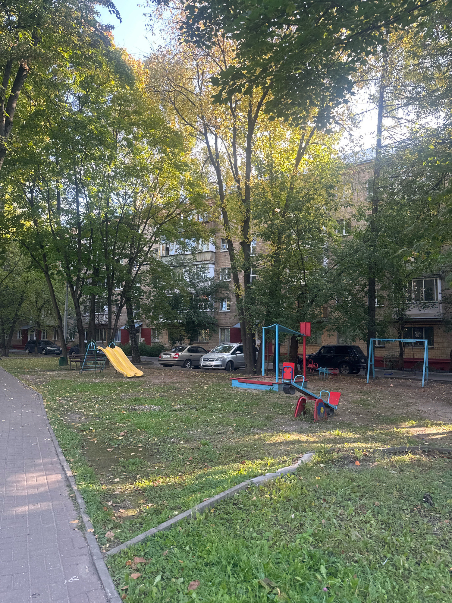 The image depicts an outdoor courtyard in Balashikha, Russia, with an apartment building in the background. A paved walkway runs along the left side of the frame, bordered by grass and fallen leaves. In the center of the courtyard, there is a children's playground area with a yellow slide, a sandbox with blue and red trim, a seesaw with red and blue accents, and a swing set with blue frames. Several cars are parked in the background, and trees with green and yellowing leaves are scattered throughout the scene, suggesting it is autumn. The overall impression is a residential courtyard in a Russian city.
