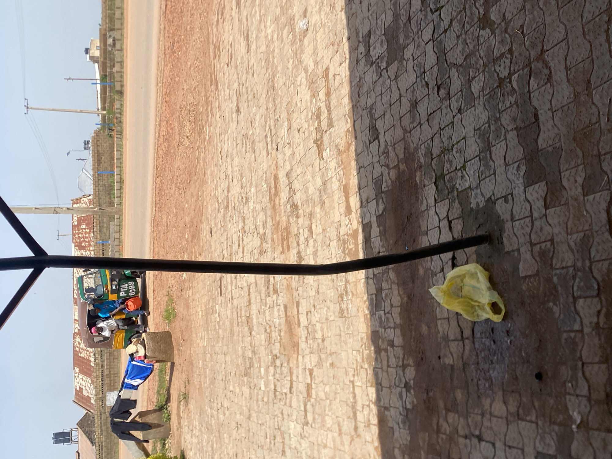 This is a full, eye-level, outdoor shot taken in Kwang, Nigeria, with a focus on a paved area in the foreground and a road and buildings in the background. A black metal pole, part of a structure, runs vertically through the right side of the frame, casting a shadow on the textured, light brown and gray pavement. A crumpled yellow plastic bag lies on the pavement in the lower left quadrant, near a dark, wet patch and some dark debris.
In the mid-ground, a three-wheeled vehicle, commonly known as an auto-rickshaw or "keke napep," is parked. It is green and yellow with the license plate "PTA 9388" visible on the side. Two men are interacting with the vehicle. One, wearing a blue shirt and a pink headwrap, is leaning into the driver's side, holding a red bucket. Another man, in a blue shirt, is partially visible in the driver's seat. A third person, wearing a white shirt, is in the passenger seat. To the left of the vehicle, several pieces of clothing, including black pants and a blue and white shirt, are draped over a concrete block.
The background shows a dusty road, a low brick wall, and several buildings with corrugated metal roofs. Electrical poles and wires are visible against a hazy, pale blue sky. The environment appears to be a somewhat arid, urban or semi-urban setting.