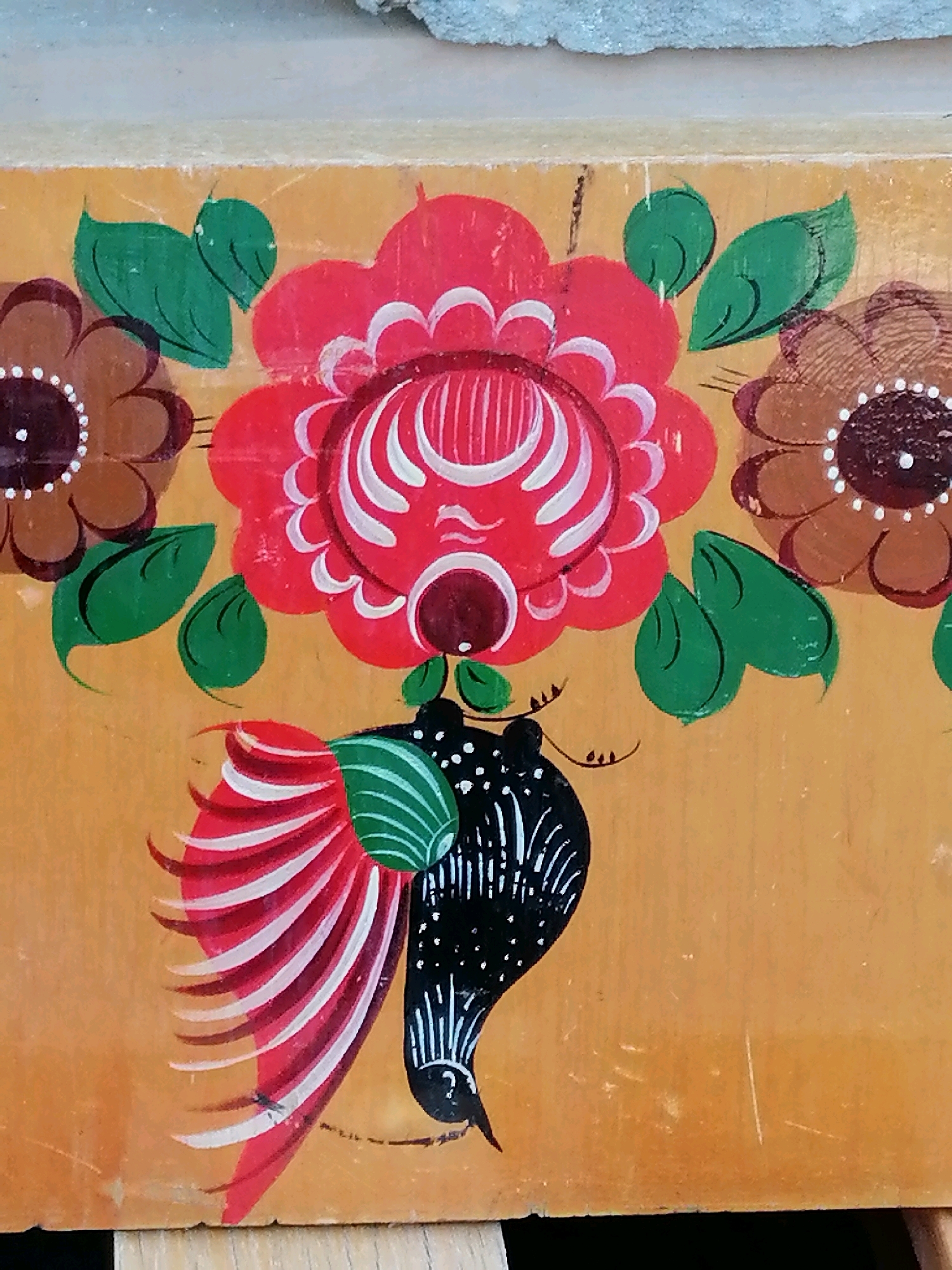 The image is a close-up, eye-level, medium shot of a painted surface. The surface is a dull, light brown, possibly wood, with visible scratches and imperfections. The painting depicts a stylized floral design and a bird. On the top half of the image are two brown flowers with darker centers and white dots. Between these flowers is a large red flower with white petal details. Green leaves are interspersed around the flowers. In the bottom half of the image, a bird is depicted in profile, facing left. The bird has a black body with white speckles and a white and red striped tail. A small black insect is visible on the red tail feathers. The painting style is folk art, characterized by bold colors and simple, decorative forms. The context of Rostov-on-Don, Russia, is not visible in the image itself.