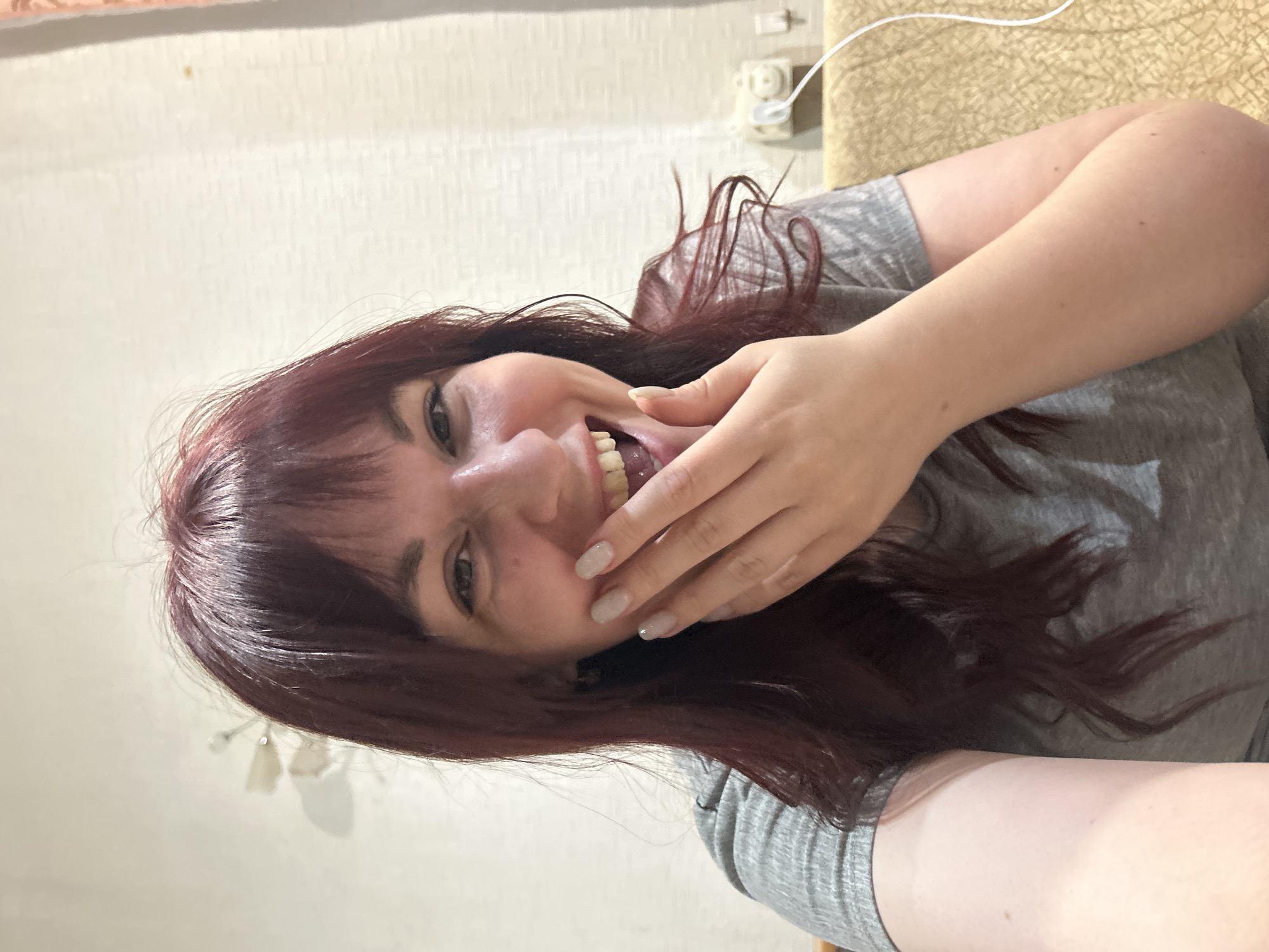 A woman with reddish-brown hair is smiling with her right hand near her face in the city of Фоки, Russia. She is wearing a gray t-shirt and has light-colored nail polish. The background is a cream-colored wall with a white electrical outlet and a beige patterned surface. The photo appears to be taken indoors.