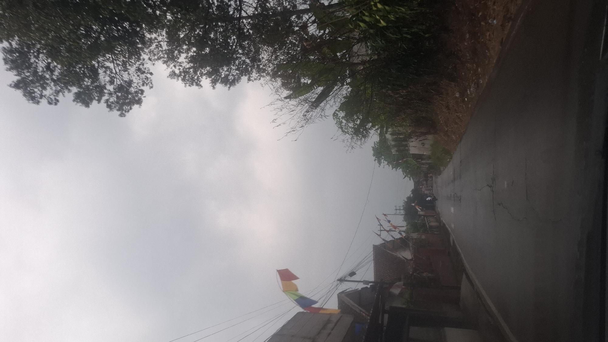 The image depicts a rural road in Sirnagalih, Indonesia, under a cloudy sky. On the left side of the road, a low-rise building is visible, with colorful flags or banners strung across wires overhead. Further down the road, more flags and houses are present. A motorcycle with two occupants is traveling away from the camera. The right side of the road is bordered by a dense cluster of trees and vegetation. The road surface appears wet and cracked. The overall impression is of a somewhat overcast day in a developing area.