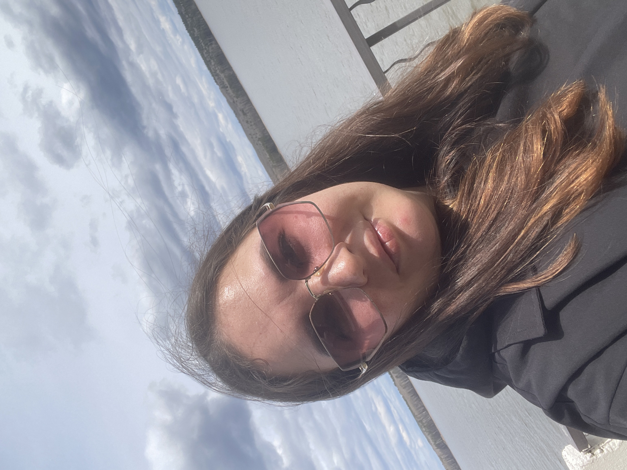 A young woman is posing for a selfie outdoors in Chaikovsky, Russia. She is wearing sunglasses and a dark-colored jacket. Her long, brown hair is partially visible. The background consists of a body of water, possibly a lake or river, with a visible shoreline and cloudy sky above. A structure of white color is visible next to her, possibly a building. The image is taken from a slightly elevated angle, as the woman is looking slightly down at the camera.