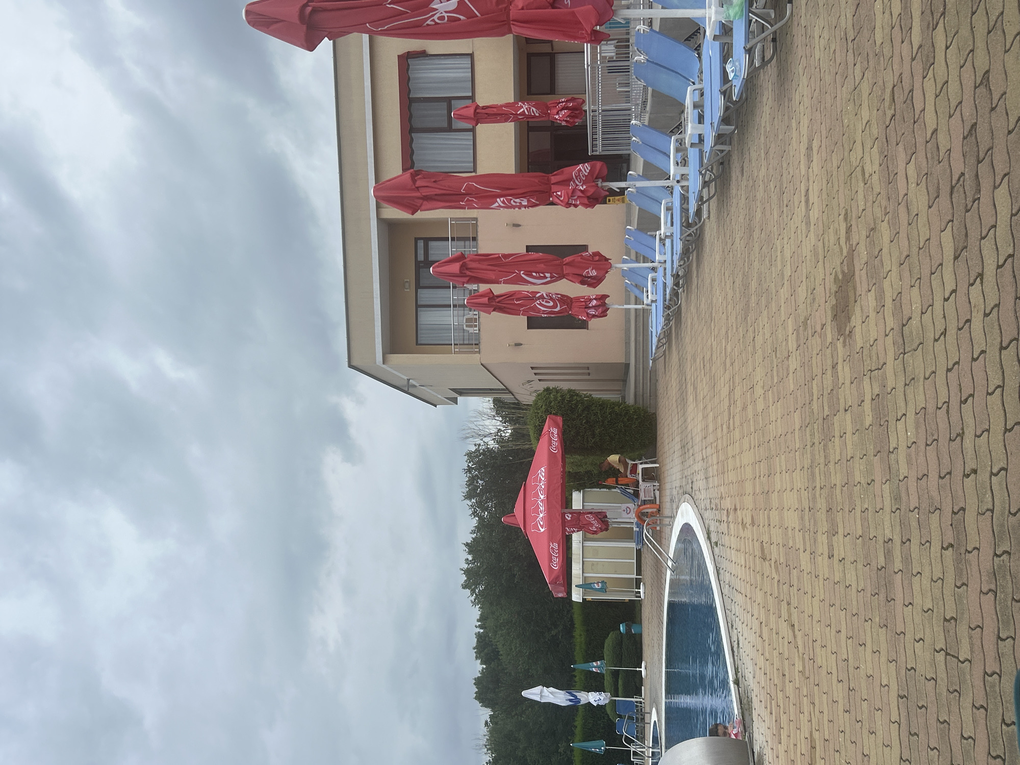 The image depicts an outdoor swimming pool area at a resort or hotel. In the foreground, a paved area with interlocking brick pavers leads to a blue swimming pool on the left. The pool has a white border and metal handrails. Some lounge chairs are visible in the pool. To the right of the pool, several closed red umbrellas with Coca-Cola branding stand on poles. Beyond the umbrellas, a beige, two-story building with windows and balconies is visible. A line of blue lounge chairs is arranged in front of the building. In the middle ground, a lifeguard sits on a white chair under a red umbrella, attending to the pool. Green bushes and trees form a backdrop behind the pool and building. The sky is overcast with grey clouds. The location is identified as Unknown, Unknown.