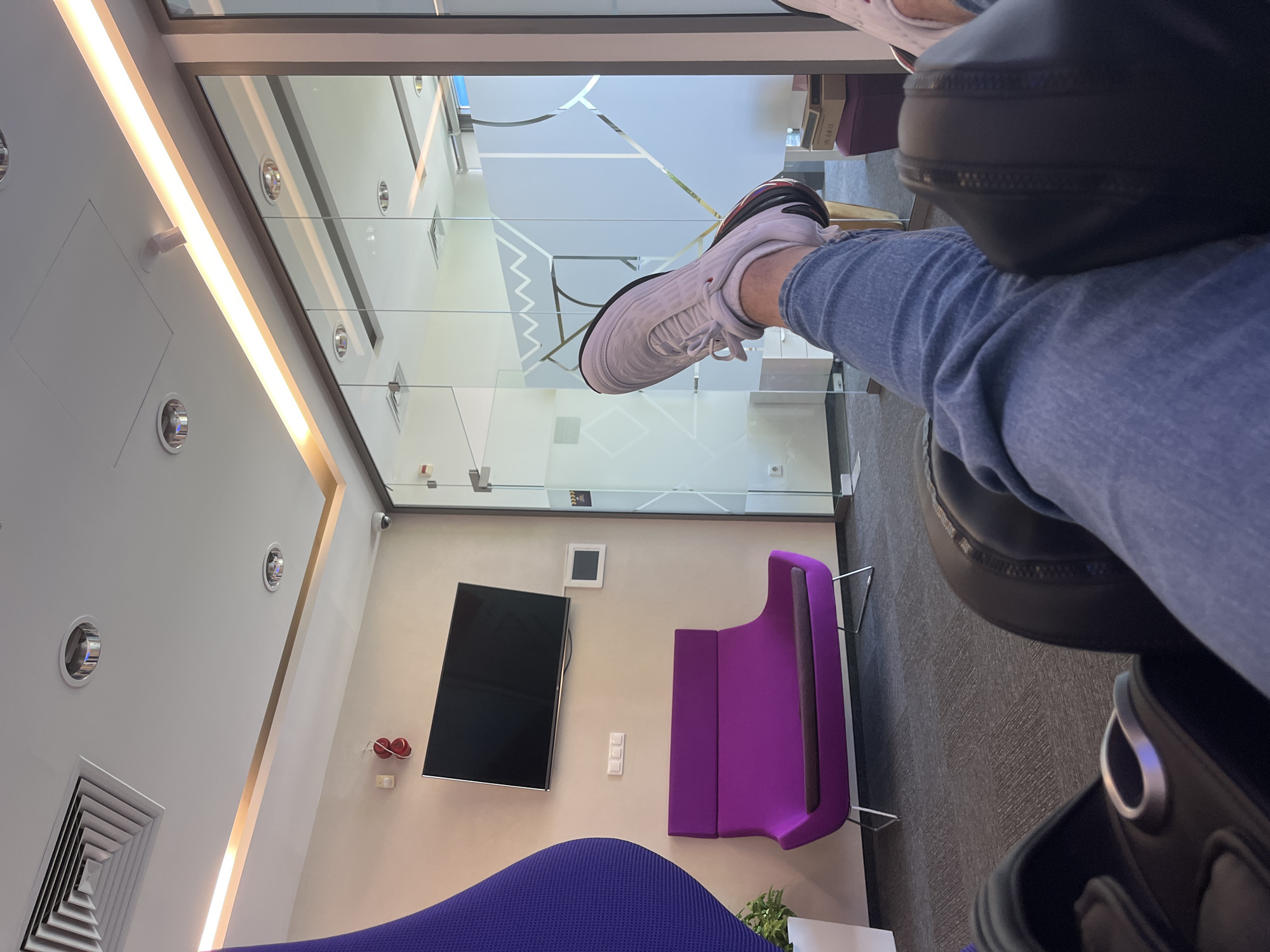 The image shows the perspective of someone sitting in a lounge or waiting area. A person's legs are extended, with one leg in a white sneaker resting on the arm of a black leather chair. The person is wearing blue jeans.
The room is modern and brightly lit with recessed lighting and a light strip along the ceiling. The walls are painted white. A large glass partition separates the seating area from another space. The glass has a geometric design etched onto it.
In the background, there is a black flat-screen TV mounted on the wall, along with a purple chair and a section of a blue couch. There are also some wall-mounted controls or electrical outlets visible near the TV. The room appears to be clean and well-maintained.