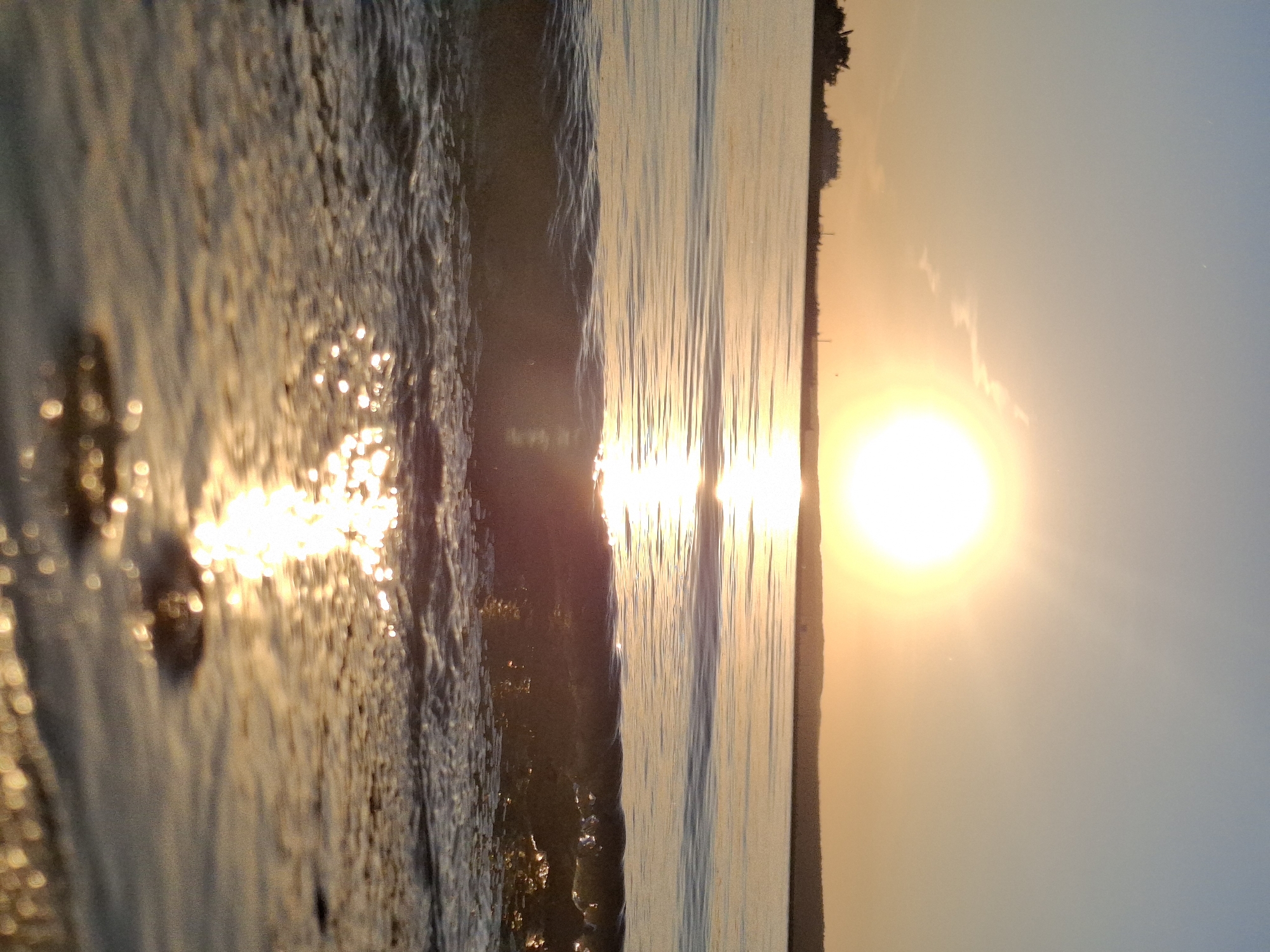 The image shows a sunset over water, likely a beach or lakefront scene. The sun is low in the sky, casting a golden glow across the water and sky. Waves are visible in the foreground, reflecting the sunlight. The location is Unknown, Unknown. The scene is serene and depicts a natural landscape.