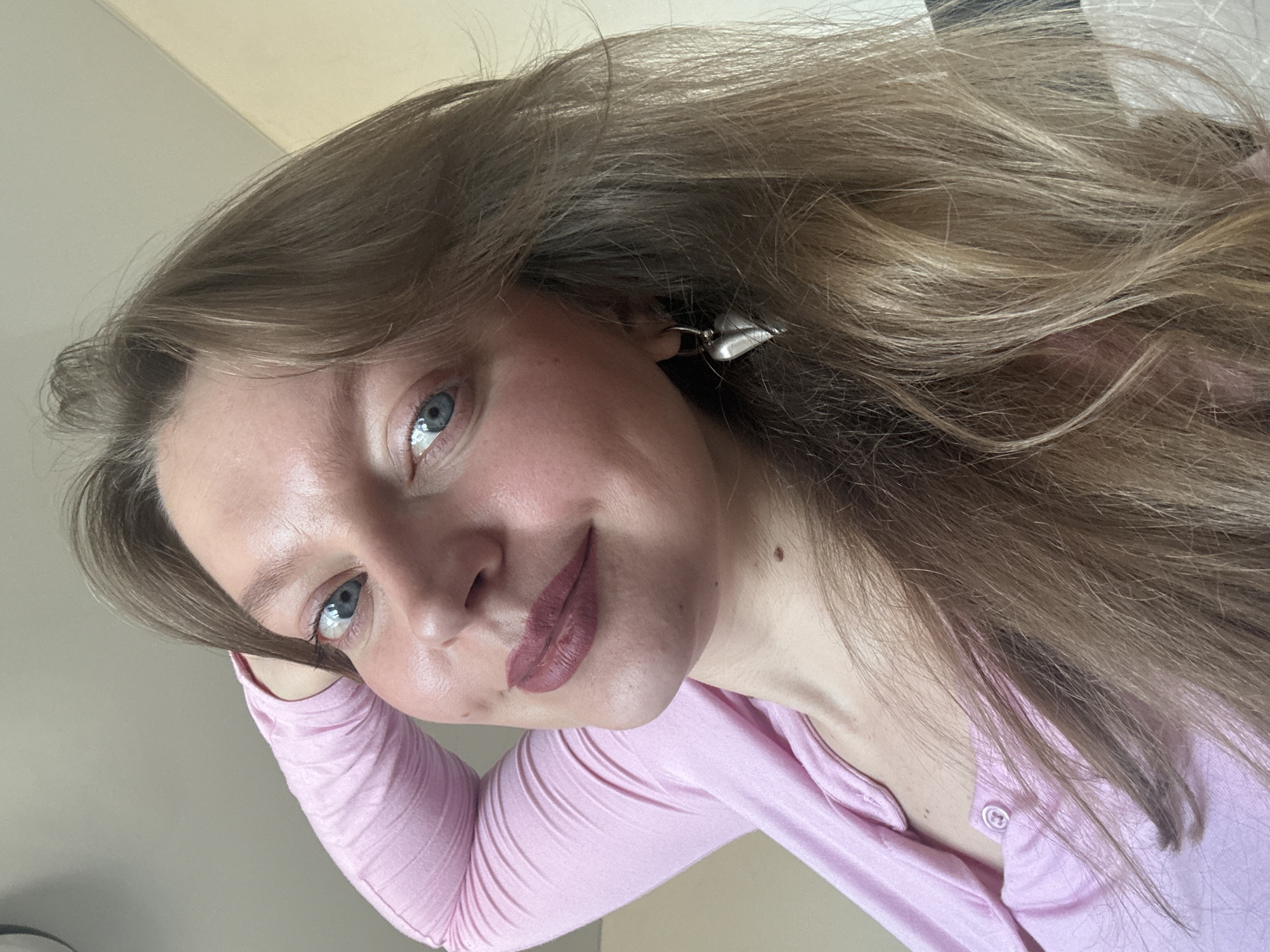 A close-up photograph of a woman with light brown, wavy hair and blue eyes, wearing a light pink, long-sleeved shirt. She has fair skin with subtle makeup, including lipstick. She is wearing a silver earring. The background appears to be a neutral-colored wall. The image is taken from a slightly elevated perspective. There is no indication of the city of Izhevsk, Russia, or any other location.