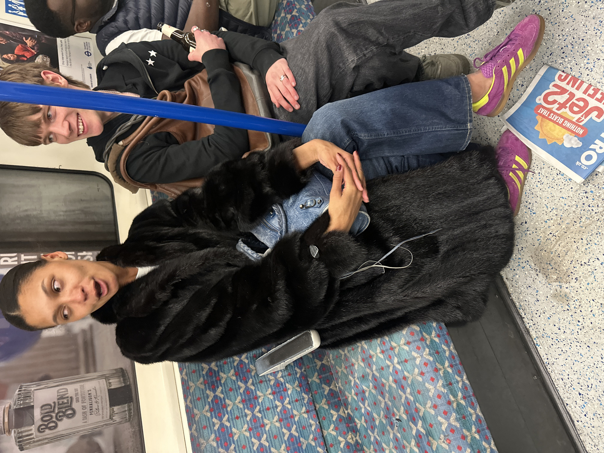 The scene depicts the interior of a London Underground train car, indicated by the distinctive patterned seating and blue vertical handrail. Three individuals are seated on a bench along the side of the carriage.
In the foreground, a woman of apparent mixed ethnicity, with dark hair pulled back, occupies the left side of the frame. She wears a long black fur coat, blue denim wide-leg jeans, and purple Adidas sneakers with yellow stripes. Her mouth is slightly open, and she looks towards her right (viewer's left). A silver-colored mobile phone rests on the seat beside her left hip. Her hands are clasped in front of her, displaying dark red nail polish, and she holds a light blue denim-textured handbag on her lap. In front of her feet on the speckled grey and blue floor, a blue and white "Jet2" advertisement or newspaper flyer is visible, stating "NOTHING BEATS THAT FEELING."
To the woman's right, a Caucasian male with light brown hair and bangs is seated. He wears a black hooded jacket with two white star emblems on the left sleeve, dark grey trousers, and has a brown leather-like satchel slung over his right shoulder. He is smiling and looking towards his left (viewer's right). His right hand holds a brown glass bottle, and a silver ring is visible on his left ring finger. A prominent blue vertical handrail separates him from the woman.
Partially visible to the far right is a third individual, appearing to be a Black male, wearing a dark puffer vest over a white garment. Only his head and upper torso are within the frame.
Behind the woman, a white advertising panel displays a bottle of "Bold Blend, Penhaligon's" fragrance. Other advertising panels are visible in the background. The seating features a repeating pattern of blue, red 'X' shapes, and small blue diamond-like shapes.