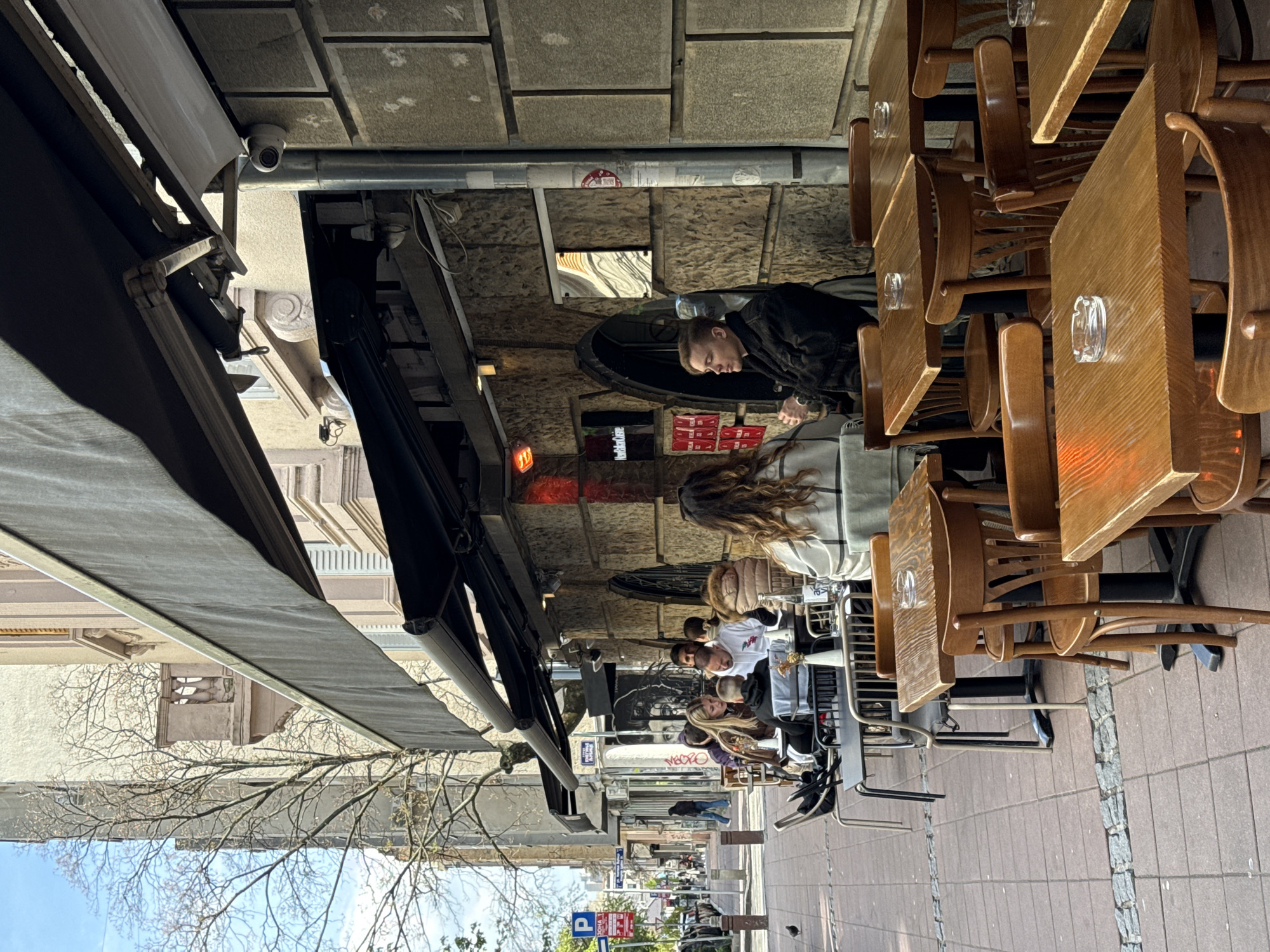 The scene depicts a street in Belgrade, Serbia with an outdoor cafe under an awning. Several people are seated at wooden tables, some with ashtrays, engaging in conversation. The sidewalk is paved with rectangular stones, and the street features typical urban elements such as parked cars, street signs, and pedestrians in the distance. The architecture along the street is varied, with stone and painted facades visible. A security camera is mounted near the awning.
