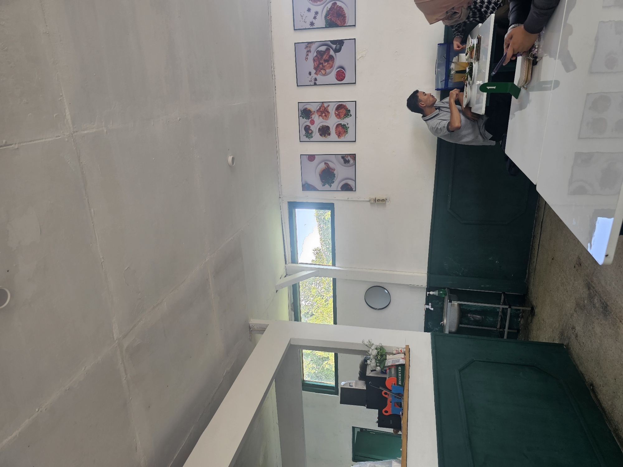 The image shows the interior of a restaurant or cafe. The walls are painted white with a dark green wainscoting. There are framed pictures of food on the wall. In the background, there are windows looking out onto trees and sky. In the foreground, there are several tables. At one table, a man in a grey polo shirt is eating, and a woman wearing a hijab is also seated at the table. Another table in the foreground is empty. There is a sink in the corner of the room. The ceiling is exposed and appears to be concrete. The floor is concrete. The overall atmosphere appears to be casual. The location is identified as Banyuurip, Indonesia, though this specific detail cannot be visually verified from the image alone.
