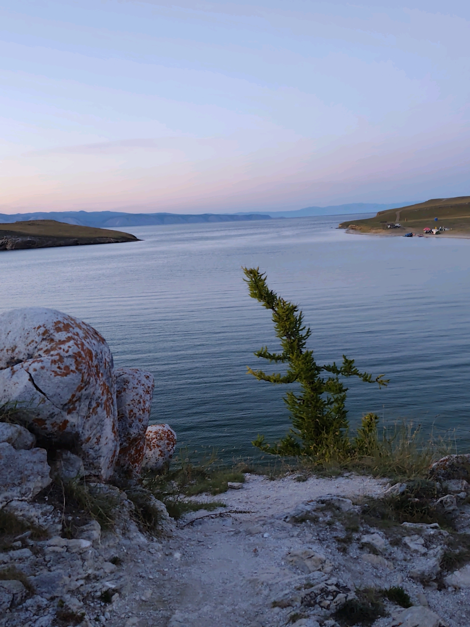 The image captures a scenic view of Olkhon Island near the settlement of Kharantsy, Russia, at twilight. The foreground features rough, rocky terrain with scattered dry grass and a large, light-colored boulder partially covered in reddish-orange lichen. A small, sparse evergreen tree with thin branches is prominently positioned to the right of the boulder, leaning out over the water.
Beyond the foreground, a vast expanse of calm, dark blue water stretches to the horizon. The water exhibits subtle ripples, indicating a gentle breeze. In the mid-ground, a gently sloping, grassy hillside forms the opposite shore, meeting the water at a sandy beach. Several vehicles, including a white truck and a few cars, are parked near the shoreline. Two small boats, possibly kayaks or canoes, are visible on the beach, along with a couple of tents, suggesting a campsite.
The background consists of a series of low, undulating hills or mountains in shades of blue and purple, receding into the distance. The sky above is a soft gradient of pale blue near the horizon, transitioning to a lighter, almost white hue higher up. Faint wisps of clouds are visible in the upper portion of the sky. The overall lighting suggests the soft light of either dawn or dusk.