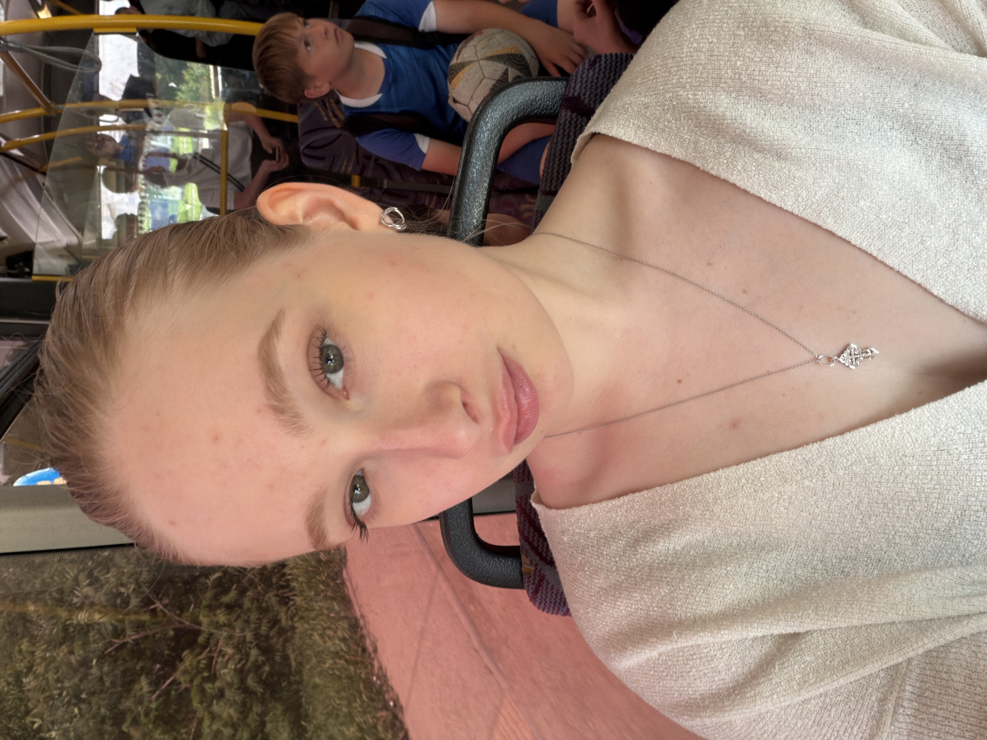The image shows a young woman on a bus in Rossiyskiy, Russia. She has fair skin, light brown hair, and is wearing a light-colored sweater and a silver necklace with a cross pendant. She is looking directly at the camera. Behind her, through the bus window, trees and foliage are visible. Further back on the bus, a young boy in a blue shirt is sitting with a soccer ball. Other passengers are visible but blurry. A black handrail is near the woman.