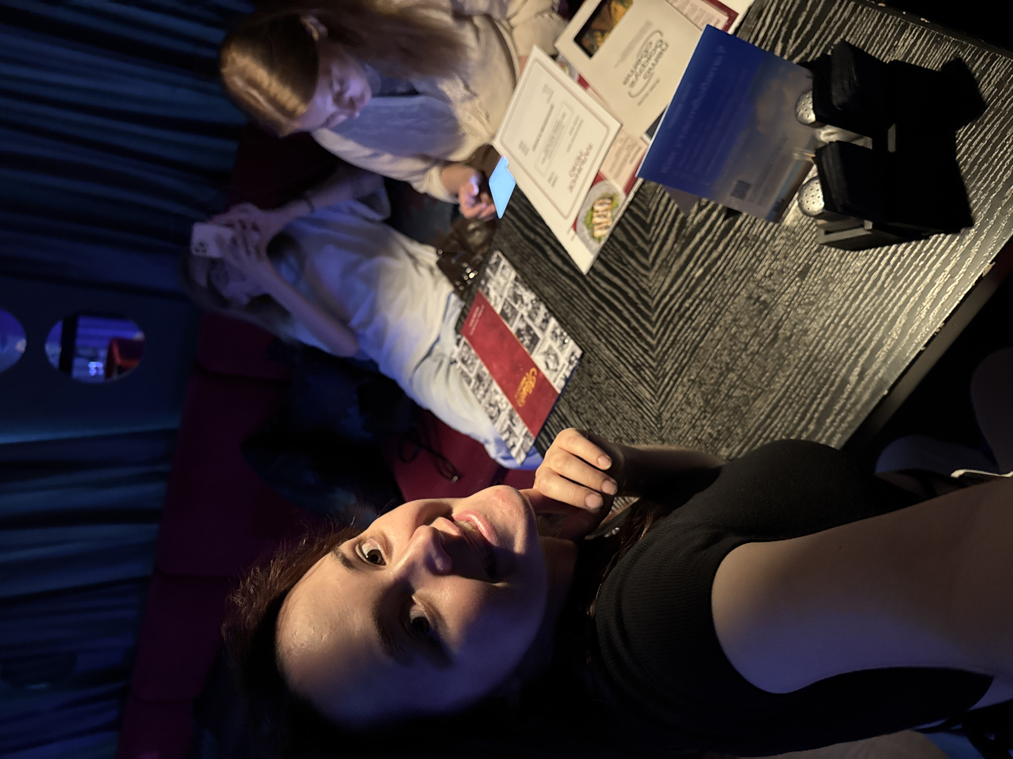 The image shows a group of three young women sitting at a table in what appears to be a dimly lit establishment, possibly a restaurant or a lounge. The woman in the foreground, on the left, is looking directly at the camera and smiling. She has long, dark brown hair and is wearing a black top. Her right hand is resting on her chin. Behind her, two other women are seated. The woman in the middle is wearing glasses and a light-colored top, and she is holding up her phone, seemingly taking a picture or recording. The woman on the right has blonde hair and is wearing a beige top, looking down at her phone.
The table they are seated at has a dark, textured surface and is cluttered with various items. There are menus or pamphlets spread across the table, some with text and images of food. A salt and pepper shaker set in a black holder is also visible. The background features a dark red sofa and dark blue curtains, with circular decorative elements visible on the wall. The lighting is dim and atmospheric, with hints of blue and red light. The overall impression is of a casual social gathering.
While the image itself does not contain specific visual cues to pinpoint its exact location within the city of Izhevsk, Russia, the context of the query suggests it is from there. Izhevsk is the capital city of the Udmurt Republic in Russia, known for its industrial heritage and cultural institutions. Without further information or visual landmarks, a more precise location within Izhevsk cannot be determined from the image alone.