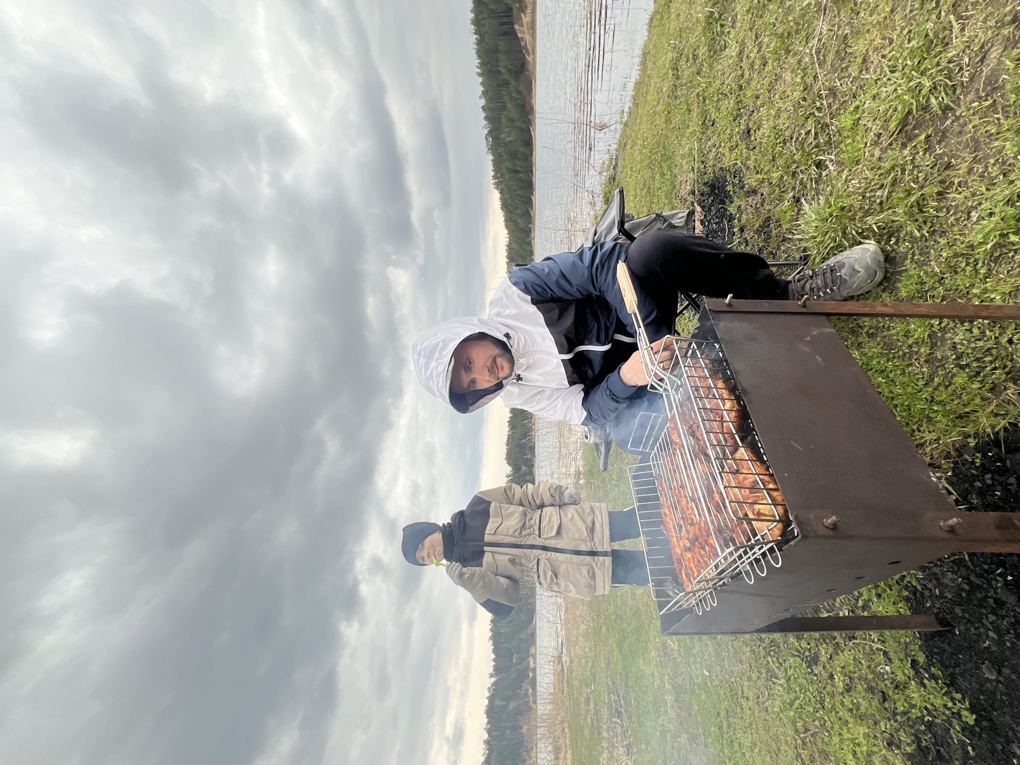 An outdoor scene depicts two individuals engaged in grilling by a lake under an overcast sky. In the foreground, a man wearing a white and dark blue hooded jacket tends to a rectangular metal barbecue grill. He holds grilling tongs and is actively cooking various pieces of meat on the grill's grate, with visible smoke rising. He looks towards the camera with a slight, friendly expression. Beside him, slightly further back, another person, dressed in a beige and black winter-style jacket and a dark beanie, stands near the grill, holding a small green object.
The setting is characterized by a grassy foreground, with patches of darker earth or ash directly around the grill. A serene lake or large body of water occupies the mid-ground, reflecting the cloudy sky and the distant landscape. The far bank of the lake is lined with a dense forest of coniferous trees. The sky above is entirely covered with thick, grey clouds, indicating an overcast day. The attire of the individuals suggests a cool or mild weather day. No discernible text is visible in the image. The overall impression is that of a leisure activity or picnic in a natural, open environment.