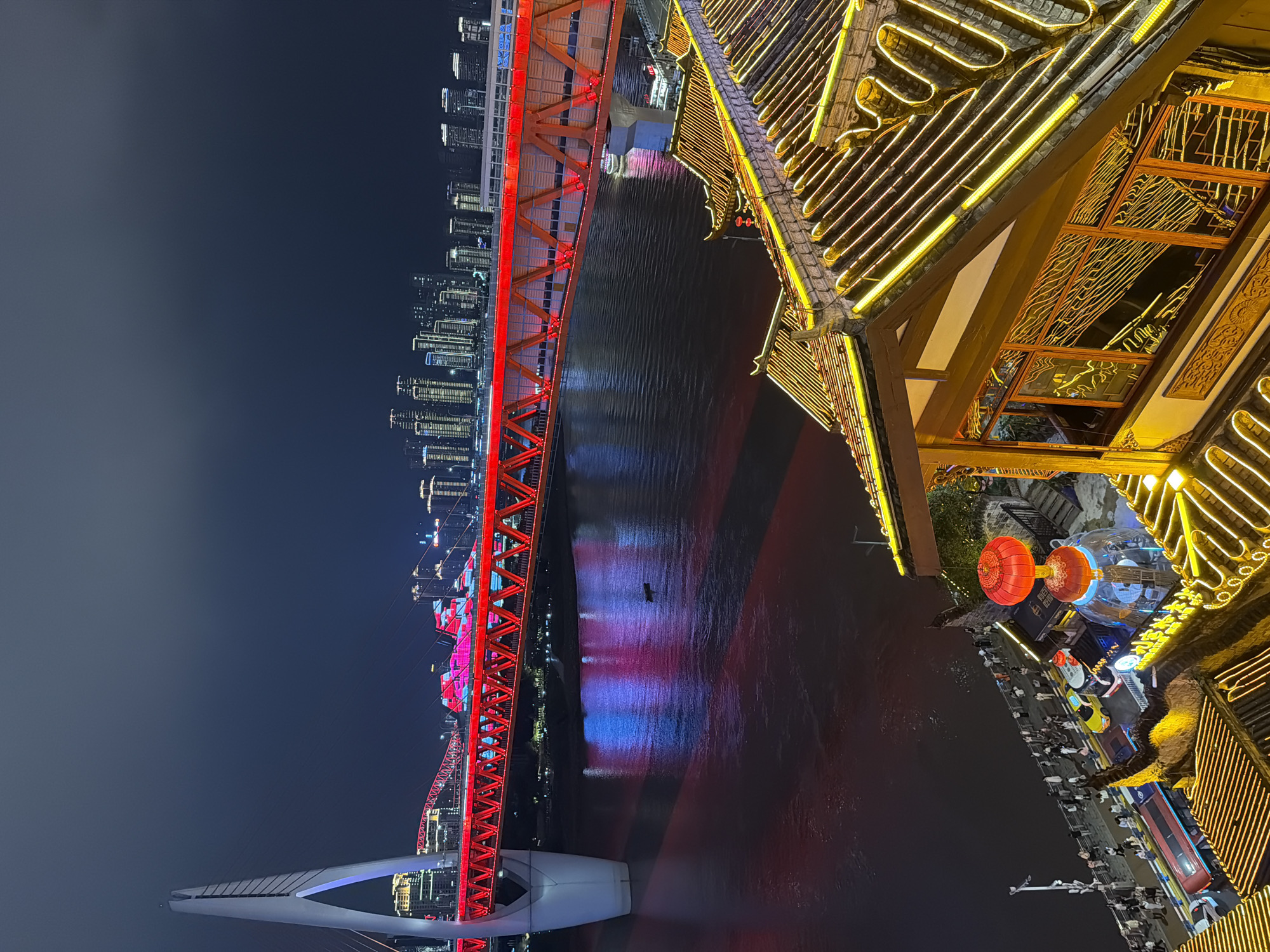 An aerial, high-angle shot captures a vibrant night scene in the Yuzhong District of Chongqing, China. A prominent cable-stayed bridge with a white, A-frame pylon spans a wide, dark river. The bridge itself is illuminated with bright red lights, casting a strong red and blue reflection onto the water. In the distance, a modern cityscape with numerous illuminated skyscrapers stretches across the horizon. In the foreground, a traditional Chinese-style building with a dark tiled roof is adorned with intricate yellow neon lights that outline its architecture. These lights also reflect in the windows of the building, creating a warm glow. A walkway along the riverbank is visible, populated by numerous individuals and several vehicles, including a red bus and a yellow taxi. Red lanterns are also a decorative element on the building in the foreground. The sky above is dark and hazy.
