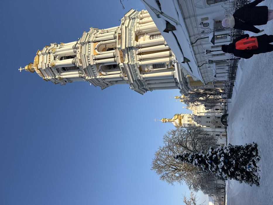The image displays a winter scene within the Kyiv Pechersk Lavra complex in Kyiv, Ukraine, under a clear blue sky. The prominent Great Lavra Bell Tower, a multi-tiered structure with white and yellow classical architecture, a golden dome, and a cross, occupies the right side of the frame. Further left, in the mid-ground, additional buildings with golden domes and white facades are visible. A low-rise white building with a steep, snow-covered roof and icicles lines the lower right foreground, separated from the path by a dark fence. The ground is uniformly covered in thick snow, and a cleared path leads from the foreground towards the background structures. To the left of this path, a snow-covered Christmas tree adorned with blue and white spherical ornaments stands near several bare deciduous trees heavily laden with snow and frost. Two individuals are walking away from the viewer on the snow-covered path: one in a dark coat with a red backpack, and the other in a dark coat and a light-colored beanie. The scene is illuminated by bright sunlight from the left.