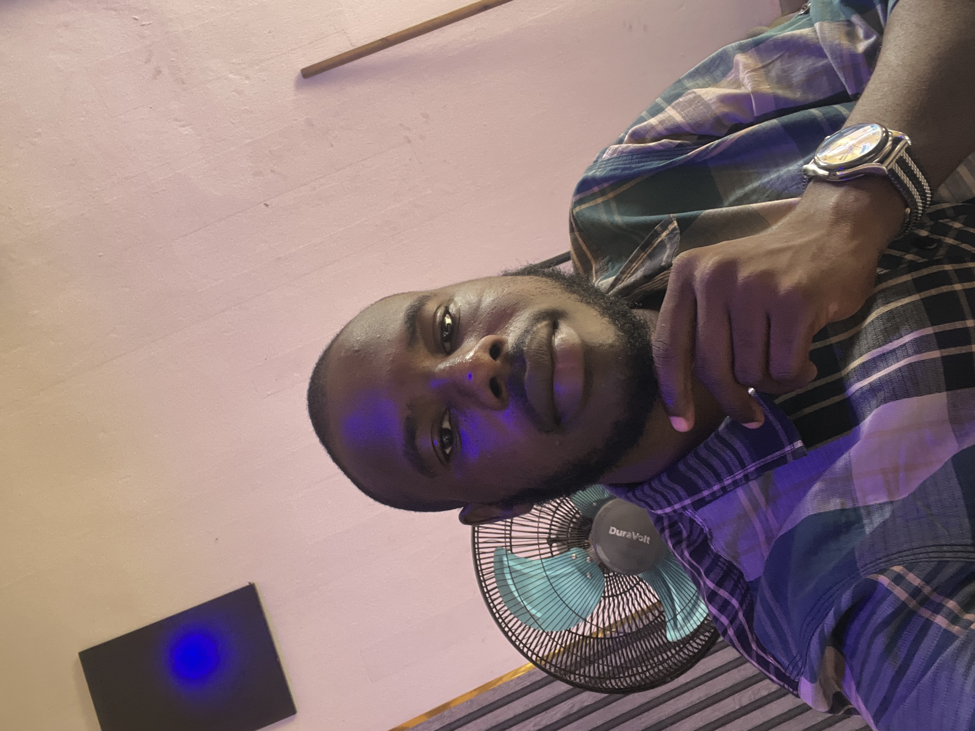 A man with dark skin, a shaved head, and a beard is looking directly at the camera. He is wearing a plaid shirt and a watch on his left wrist. His right hand is resting on his chin. Behind him, a fan with a teal-colored grill is visible. To the left of the fan, a black rectangular object with a blue light emitting from its center is mounted on a light-colored wall. The wall has a textured appearance and is illuminated by pink and purple lighting. The scene appears to be indoors, possibly in a room in Jos, Nigeria.