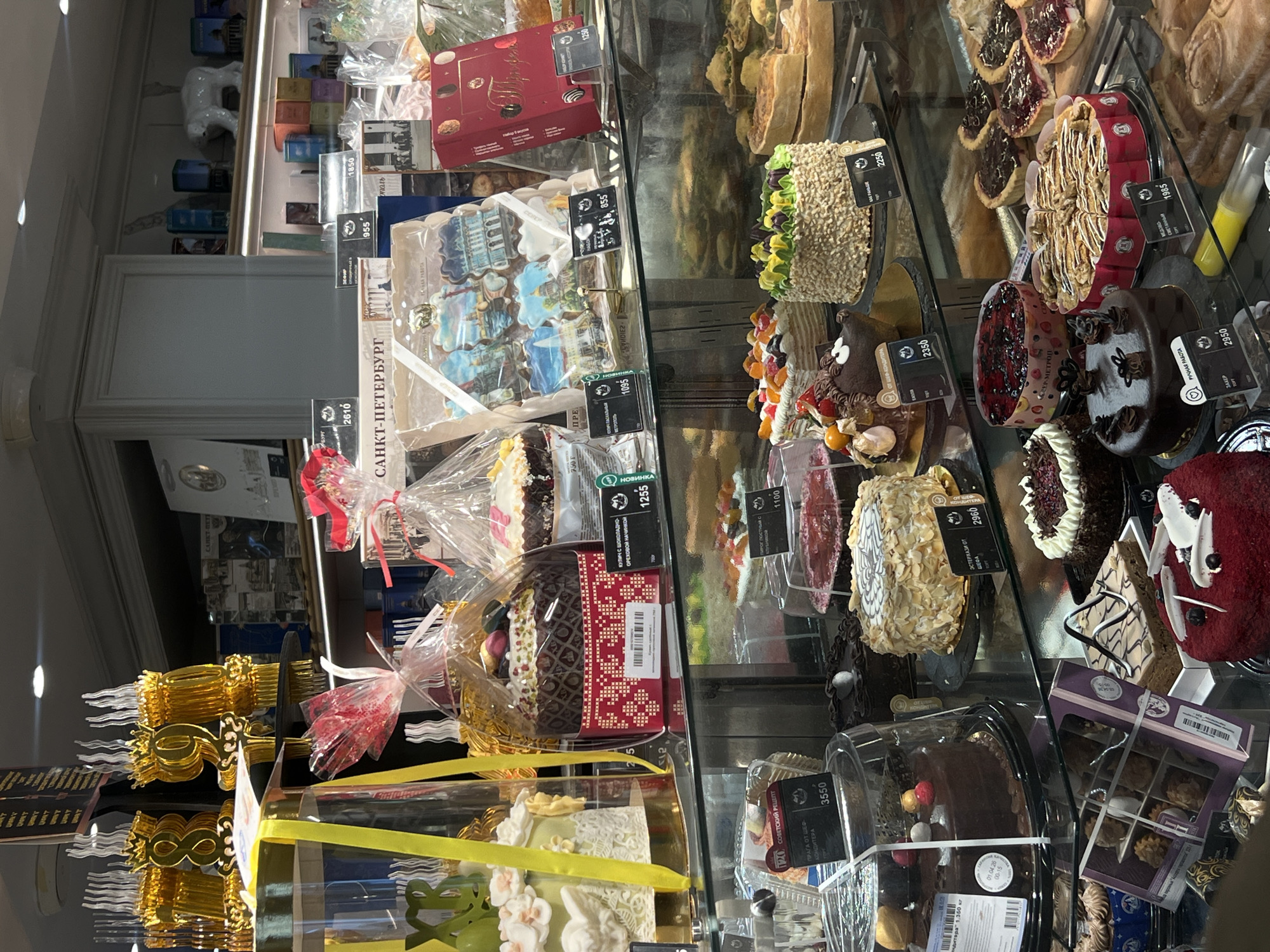 The image shows a display case in a bakery or confectionery shop, likely located in Saint Petersburg, Russia. The case contains a variety of cakes, pastries, and sweets, including elaborate cakes decorated with fruit, chocolate, and nuts. Some items are individually wrapped or packaged with ribbons. Prices are marked in Russian rubles on small tags attached to each item. The items include such desserts as "Praga cake", "Esterhazy" cake, and a special cake with a chocolate-nut filling for Easter. Other goods include various cookies and tarts. A box of sweets with the "Saint Petersburg" logo is visible. The background suggests a well-lit interior with other items displayed on shelves. The overall setting suggests a retail environment focused on selling high-end pastries and confectionery.