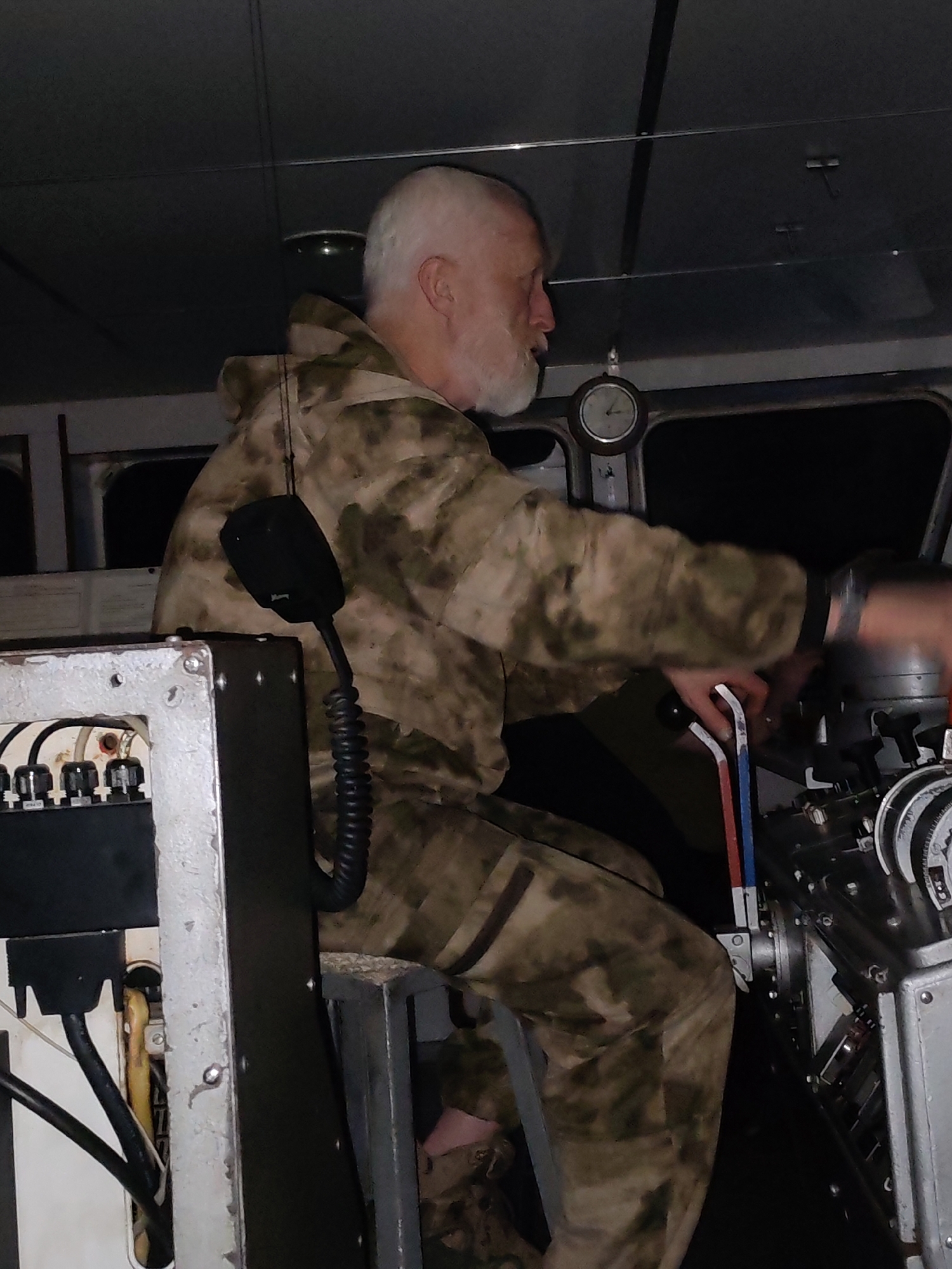 A man with gray hair and a beard, wearing a camouflage hoodie and pants, is operating machinery inside a metallic gray structure. He is seated on a stool, with his hands on levers and knobs. A microphone is attached to the structure on his left. A clock is visible in the background. The location is Zaprudnya, Russia.