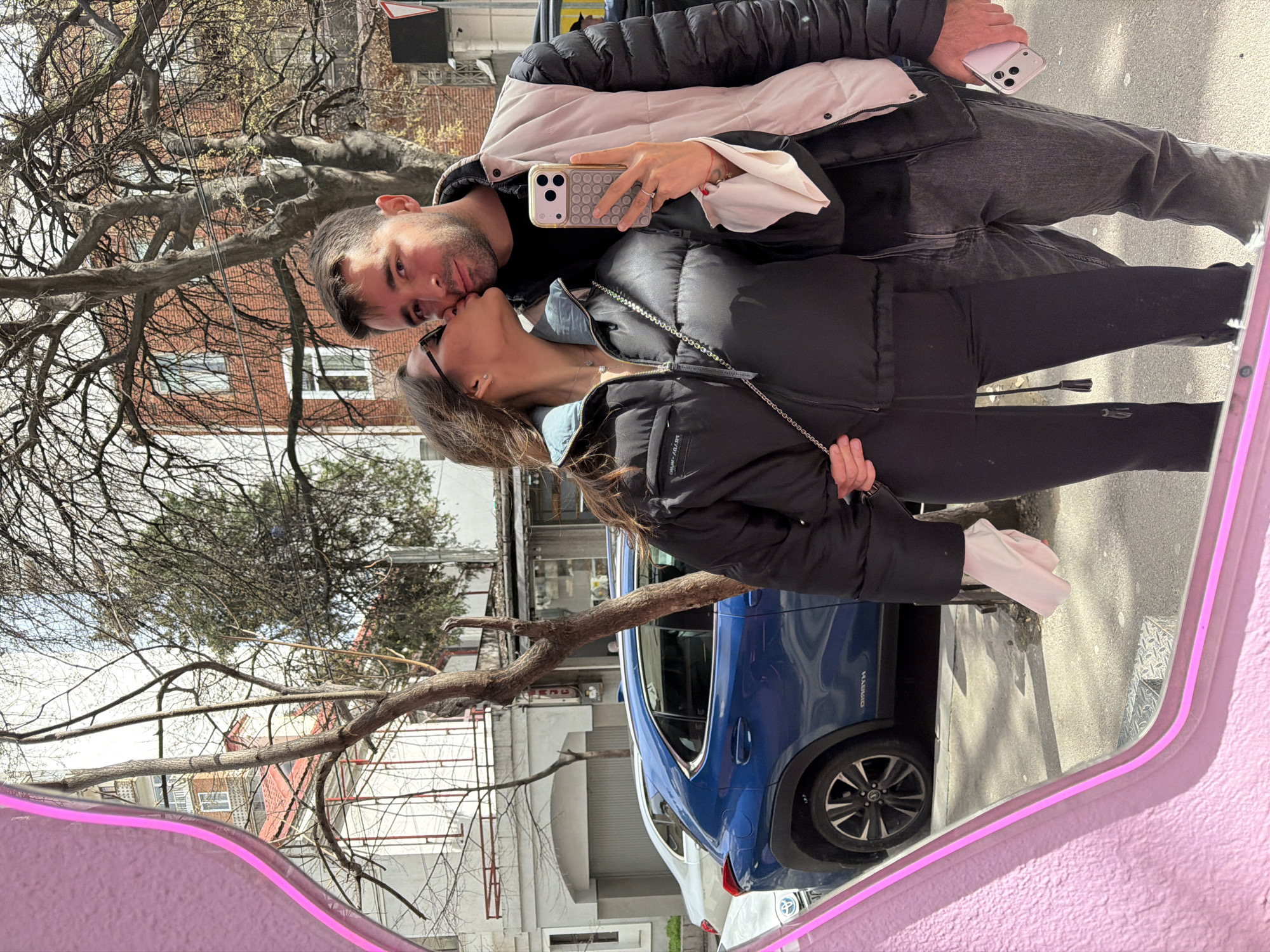 The image depicts a couple kissing, reflected in a pink-framed mirror on a city street. The male figure wears a black and white jacket, grey jeans, and holds a phone in his right hand. The female figure is dressed in a black puffer jacket and black leggings. A blue SUV labeled "Hybrid" is parked in the background. The location is suggested to be in Tbilisi, Georgia, based on the architectural style of the surrounding buildings and the type of parked vehicle. Deciduous trees with bare branches are also visible.